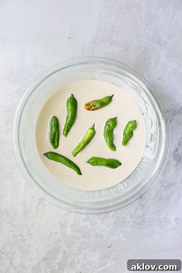 Shishito peppers being carefully dipped into the beer batter in a glass bowl.