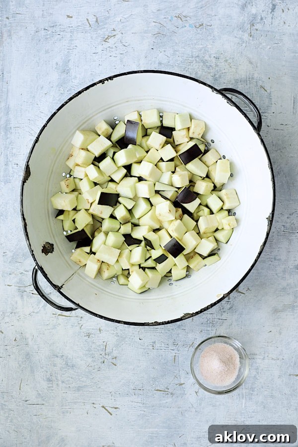 Photo of cubed eggplant generously salted in a white distressed colander, prepared for draining.