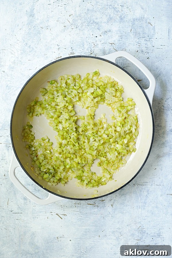 Photo of onion and celery being cooked in a white cast iron skillet, softening in olive oil.