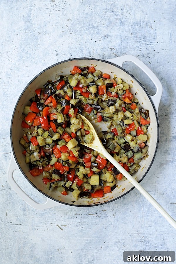 Photo of fresh ingredients for caponata being cooked together in a white cast iron skillet, including bell peppers, tomatoes, and spices.