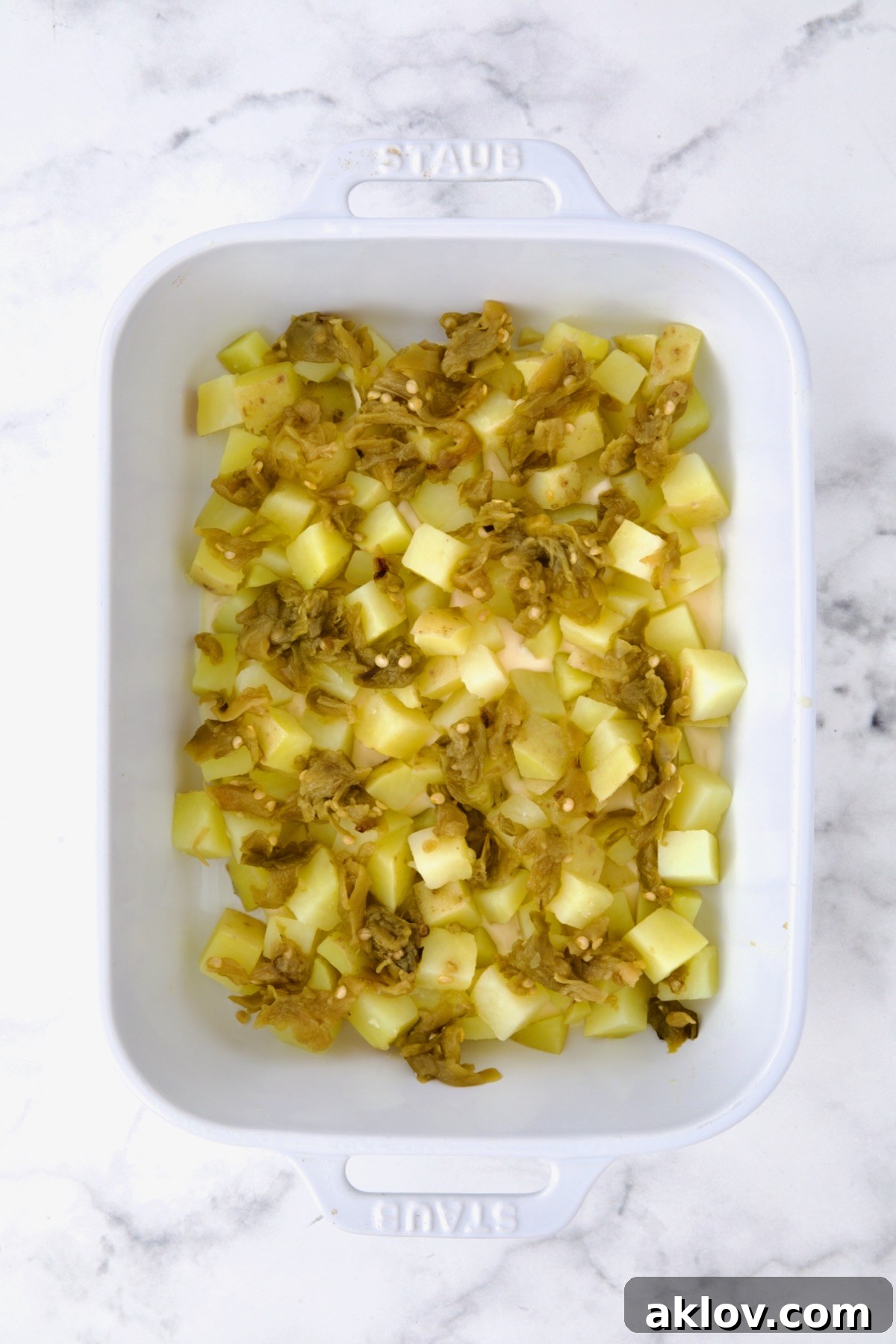 The first foundational layer of par-cooked potato cubes and vibrant green chiles spread evenly across the bottom of a white rectangular baking dish. This forms the base for the cheesy au gratin.