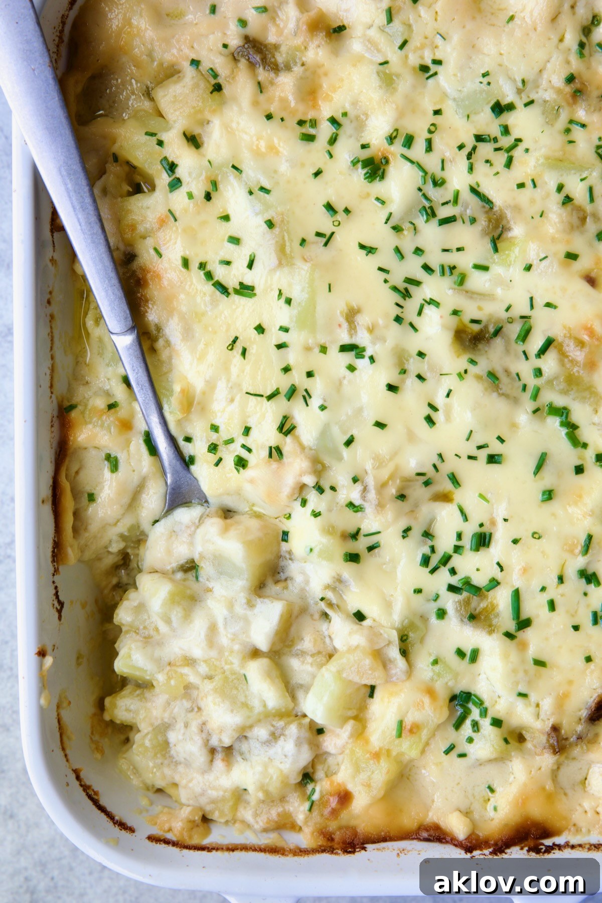 A serving of Hatch Green Chile au Gratin Potatoes being scooped from a white rectangular baking dish. The rich, cheesy layers and tender potato cubes, speckled with vibrant green chiles, are clearly visible, promising a comforting and flavorful experience.