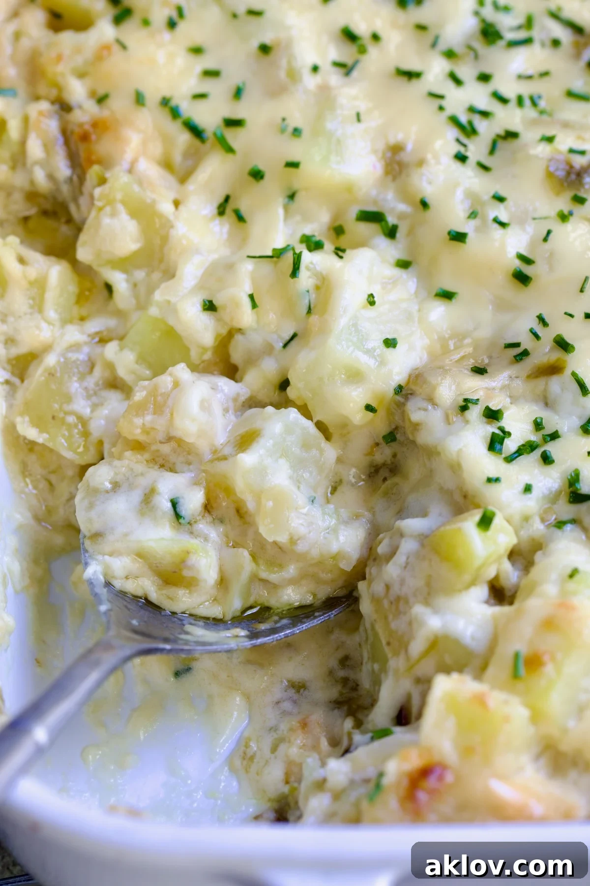 A mouthwatering close-up of Green Chile au Gratin Potatoes in a white rectangular baking dish, showcasing the golden-brown crust and a creamy, bubbling interior. A serving spoon has just scooped a portion, revealing the layers of tender potatoes and vibrant green chiles.