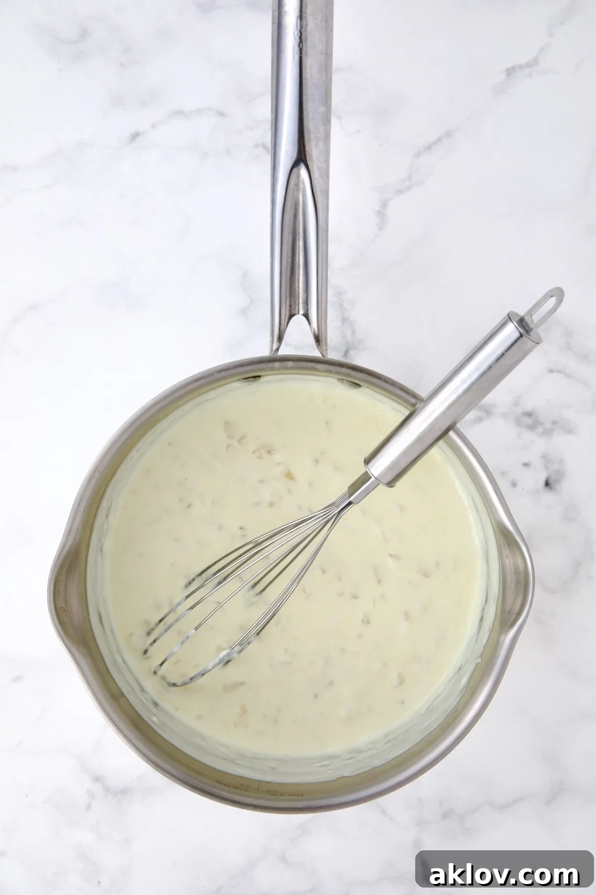 Half-and-half being slowly poured into the saucepan containing the cooked onions and flour (roux). A whisk is used continuously to incorporate the dairy, ensuring a smooth, lump-free base for the cheese sauce.