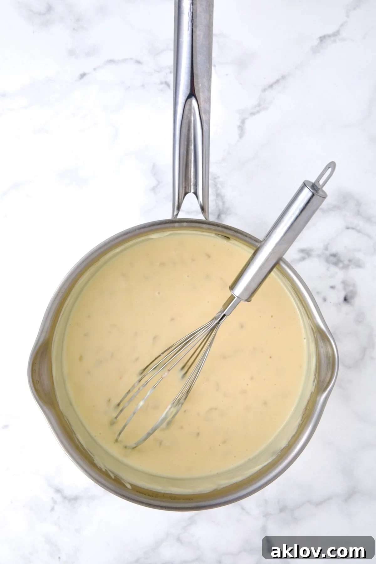 A rich, velvety cheese sauce, perfectly thickened, shown in a stainless steel saucepan with a wire whisk. The melted Monterey Jack and sharp Cheddar cheeses have created a smooth, creamy consistency, ready to be combined with the potatoes.