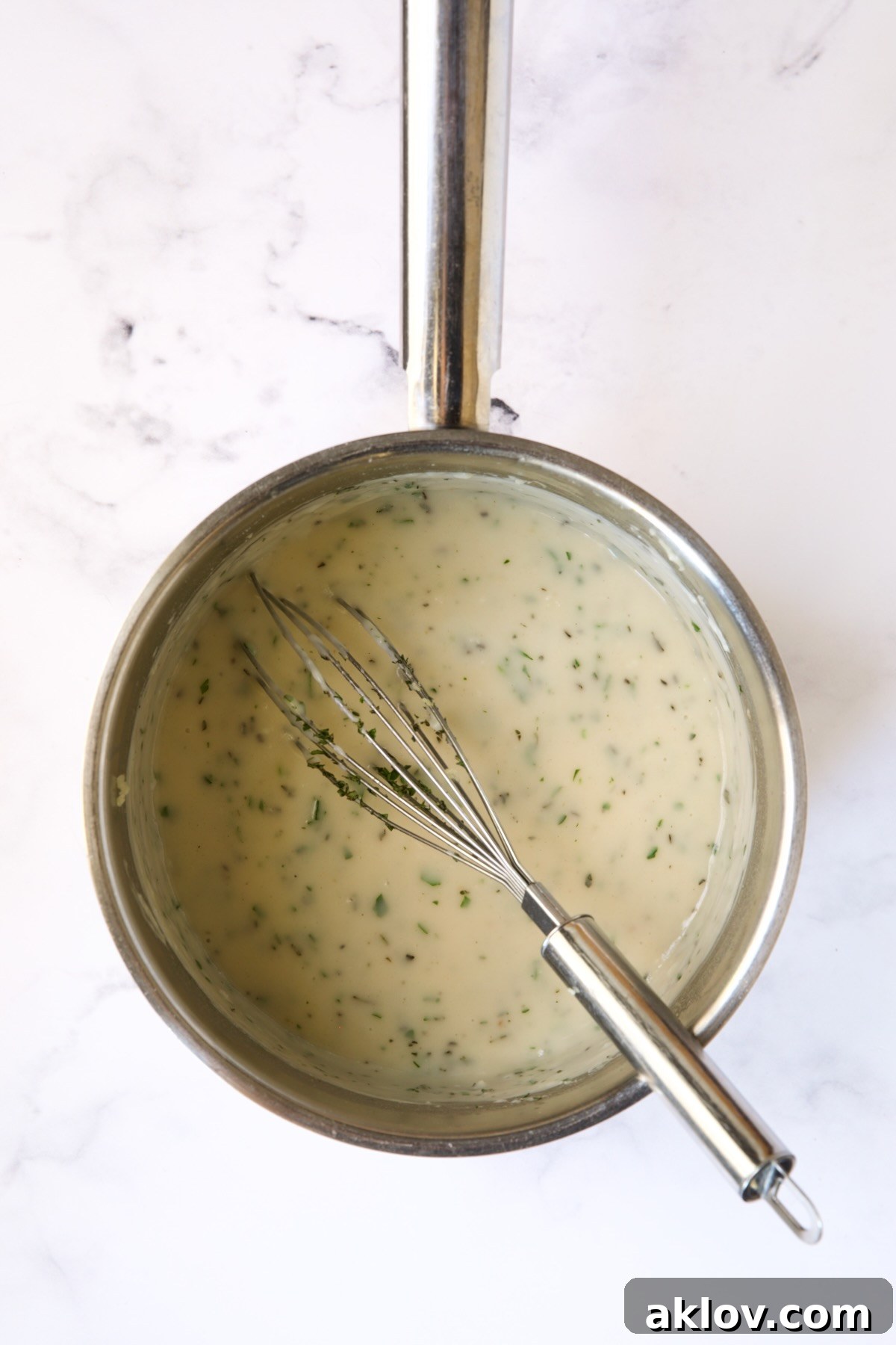 A whisk stirring the finished garlic herb sauce in a stainless steel saucepan, showcasing its smooth consistency.