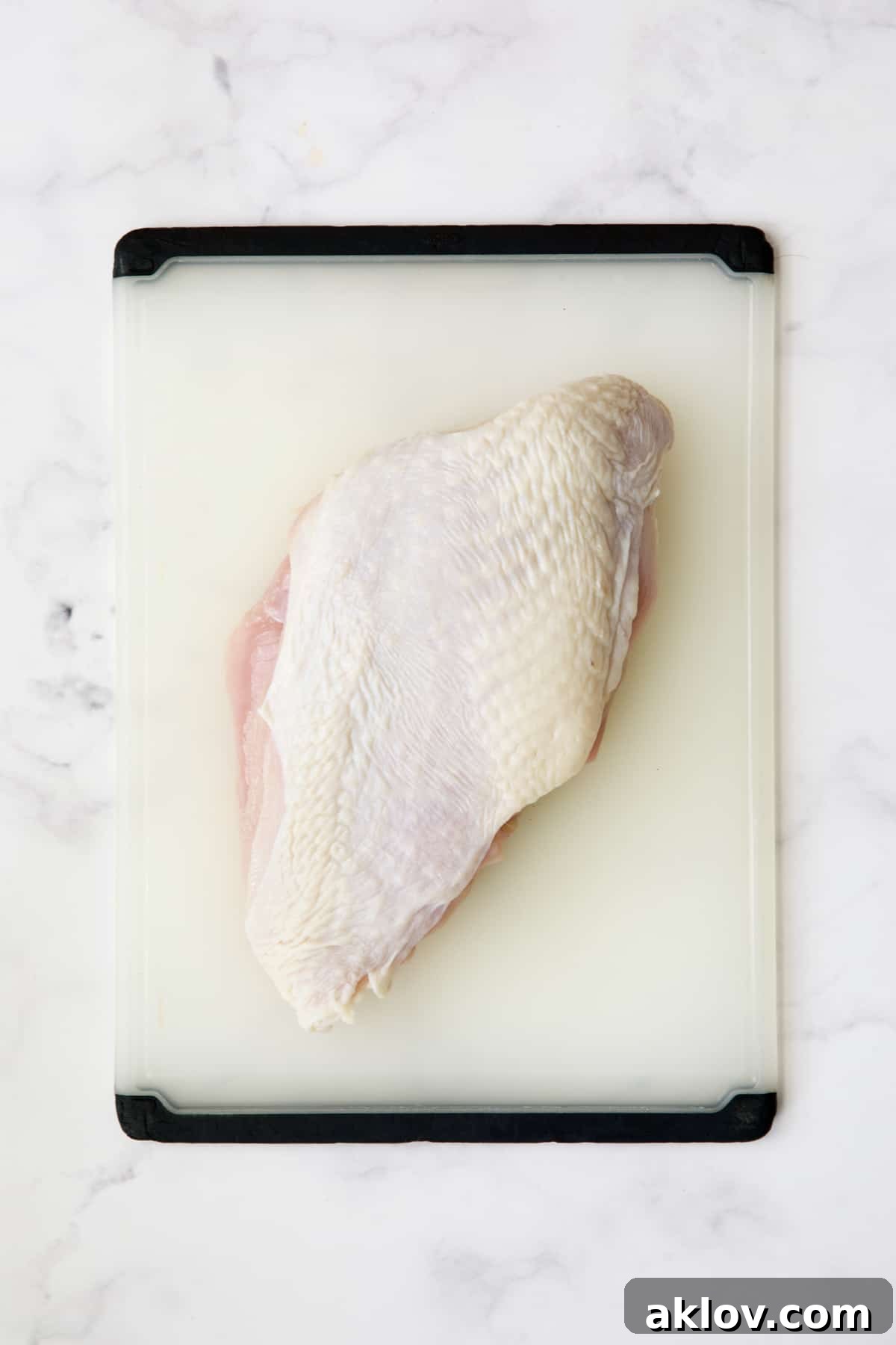 Boneless turkey breast half with skin resting on a white plastic cutting board, pre-butterflying.