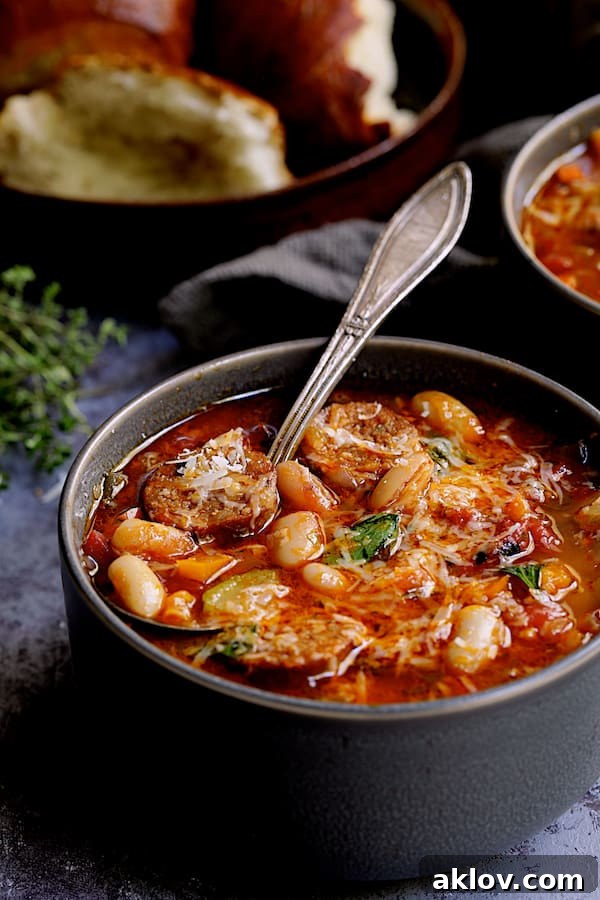 Rustic Chorizo, White Bean and Vegetable Stew 12 A close-up of a single bowl of Chorizo White Bean and Vegetable Soup, a spoon resting inside, with bread slices blurred in the background, emphasizing its deliciousness.