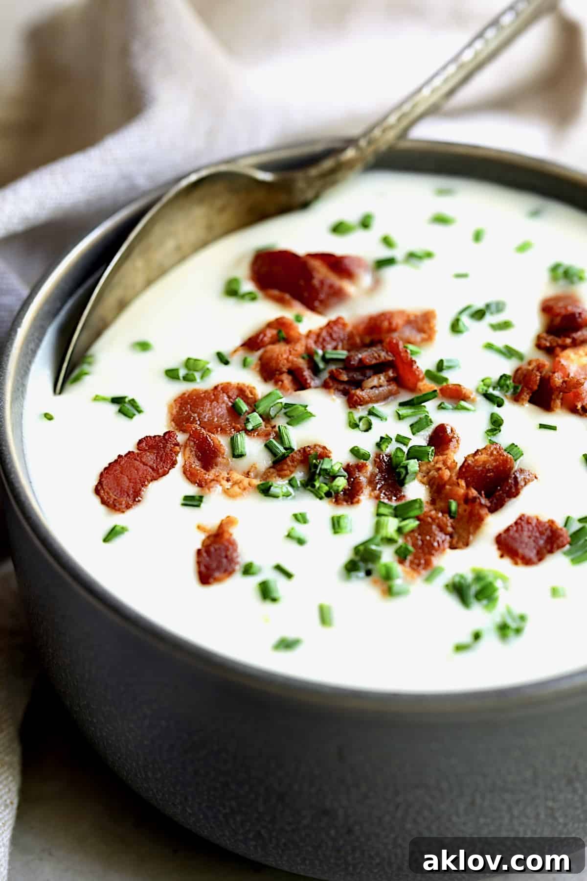 A close-up view of a single bowl of rich Potato Leek Soup with White Cheddar, elegantly garnished with crisp bacon bits and fresh green chives, showcasing its smooth, creamy texture.