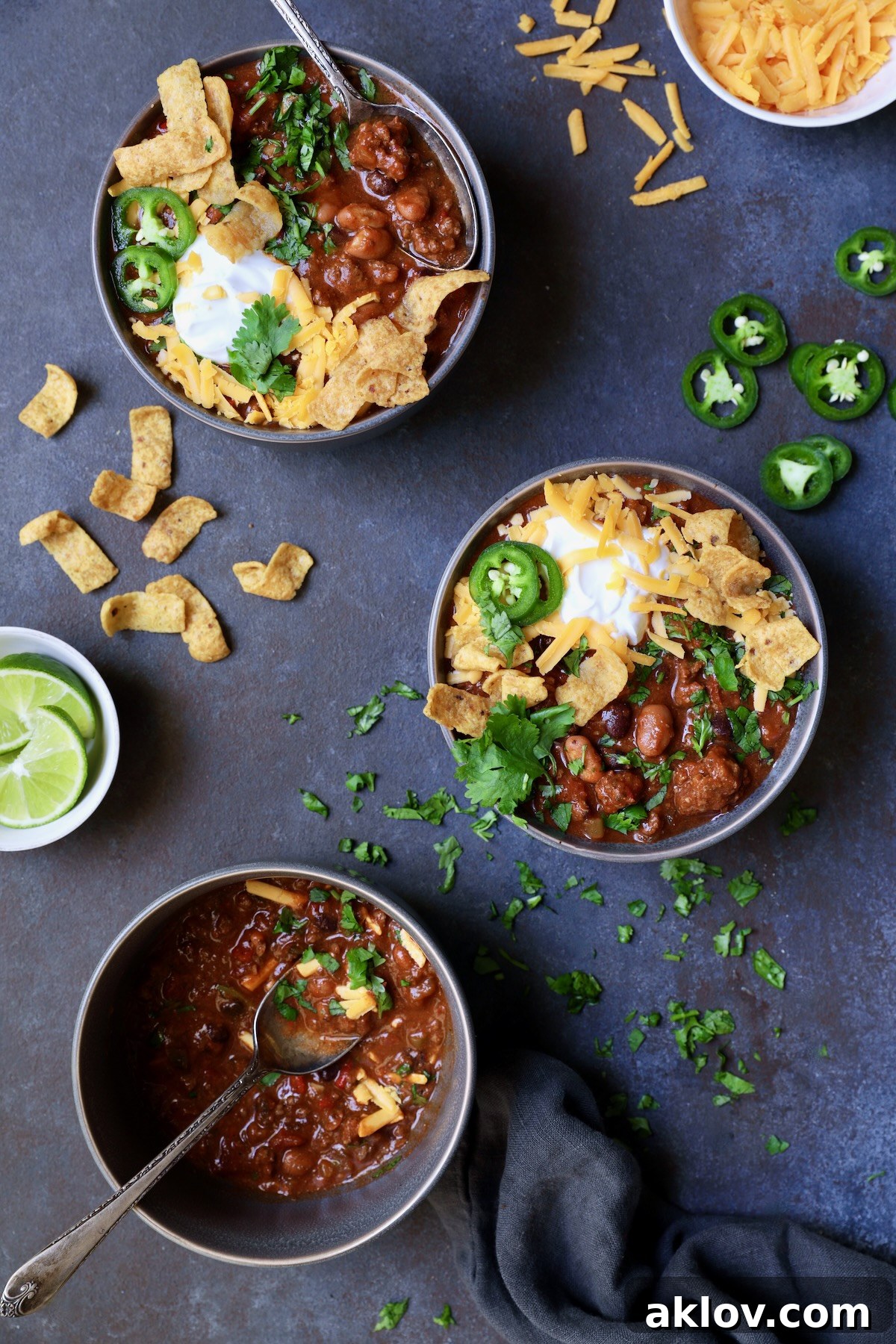 Three bowls of Game Day Chili with corn chips, cheese, sour cream jalapeno slices and cilantro in gray bowl.