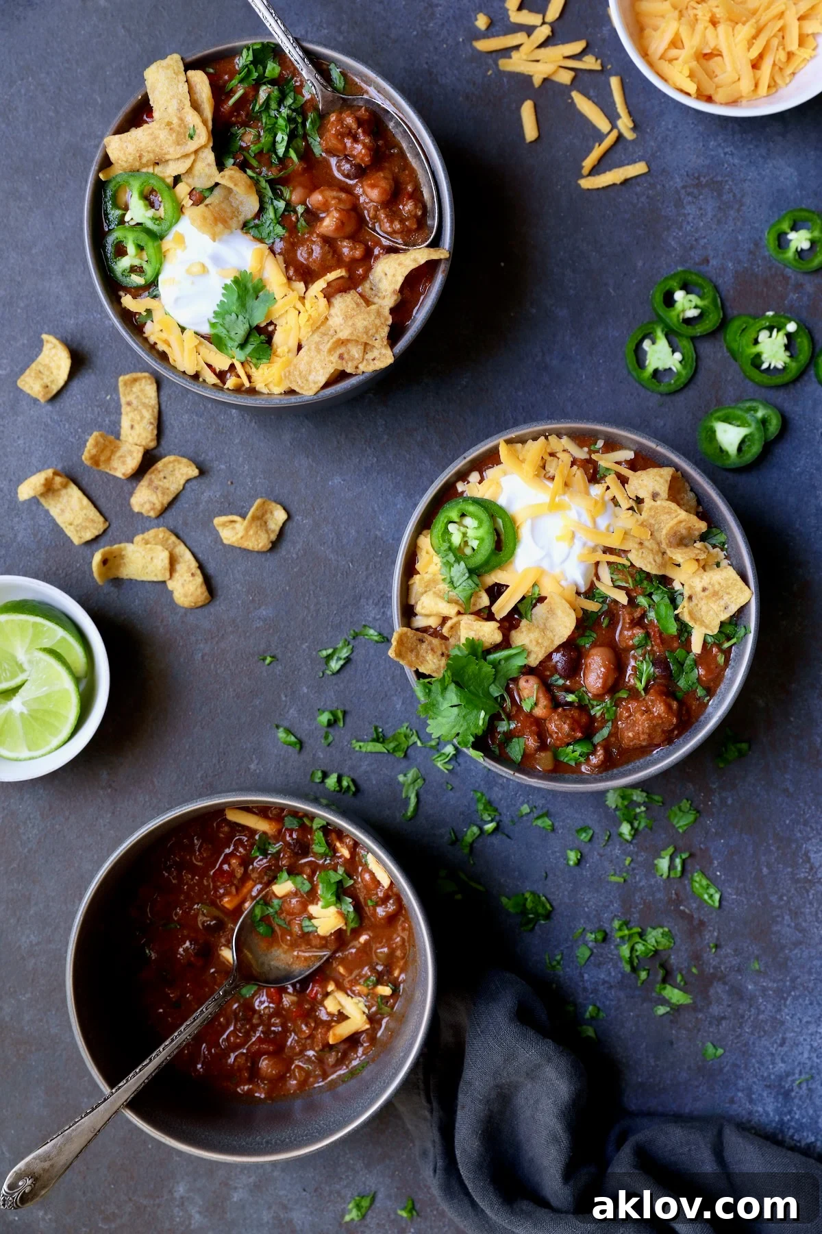 Three bowls of Game Day Chili with corn chips, cheese, sour cream jalapeno slices and cilantro in gray bowl.