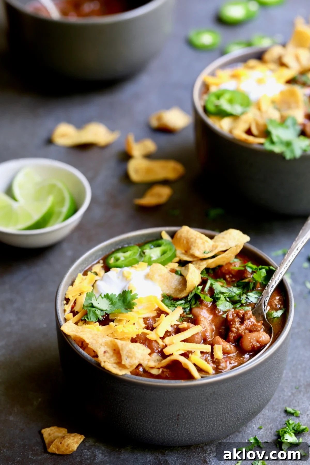 Bowls of Game Day Chili with corn chips, cheese, sour cream jalapeno slices and cilantro in gray bowl.