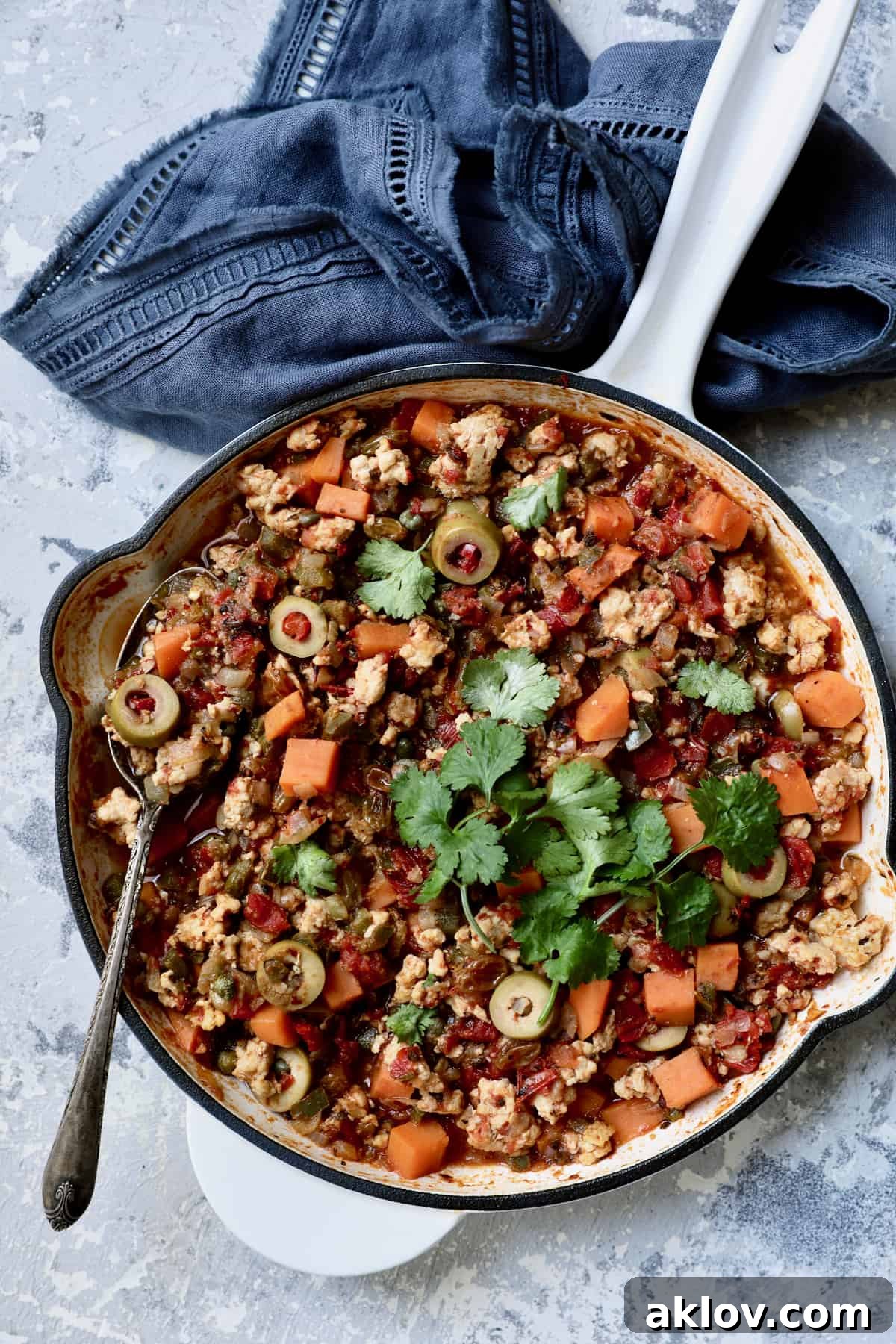 Cuban-Style Turkey Picadillo with Sweet Potatoes in white cast iron enameled skillet on blue background with blue napkin.