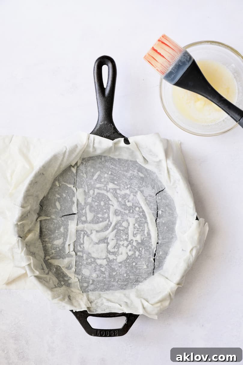 Skillet Spanakopita with Spinach and Feta 9 Photo of one piece of phyllo dough being buttered and placed in a cast iron skillet, partially covering the bottom. This illustrates the start of the layering process.