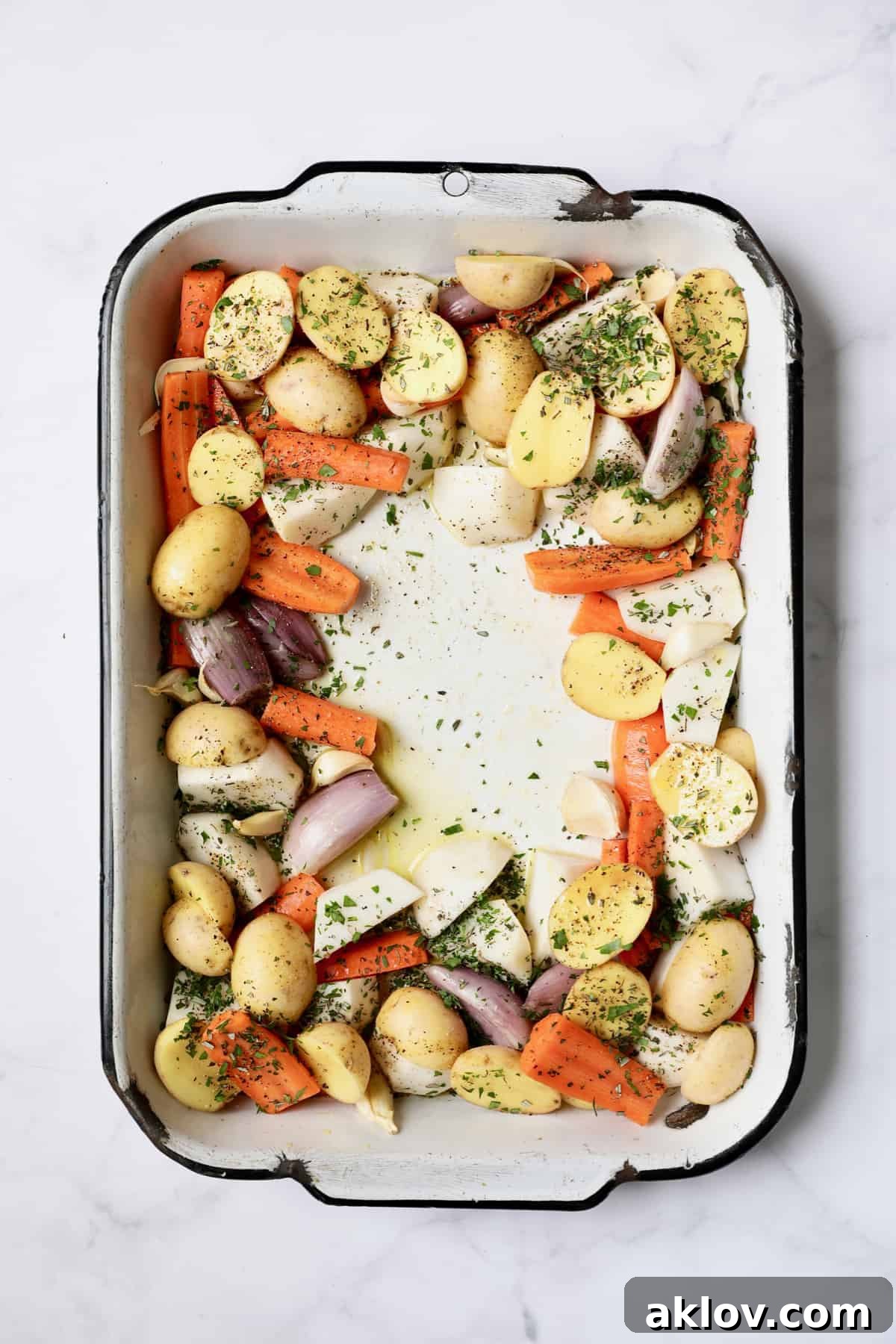 Photo of uncooked root vegetables in white roasting pan sprinkled with fresh herbs, awaiting the chicken.