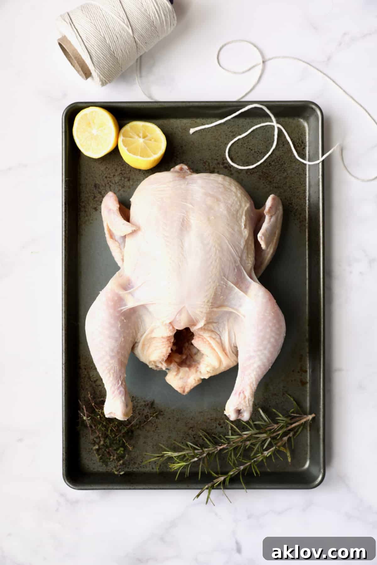 Photo of uncooked whole chicken on baking sheet with butcher's twine, lemon halves and herb sprigs, demonstrating the trussing and cavity stuffing before butter application.