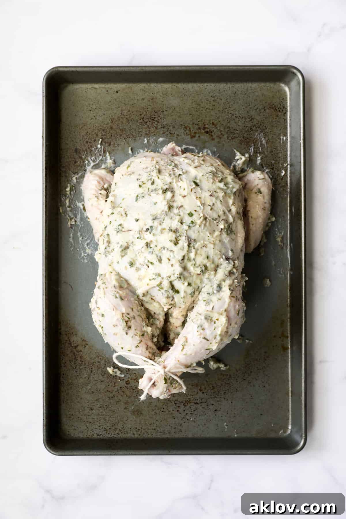 Photo of uncooked whole chicken rubbed with butter on baking sheet, showcasing the even coating of herb butter over the skin.