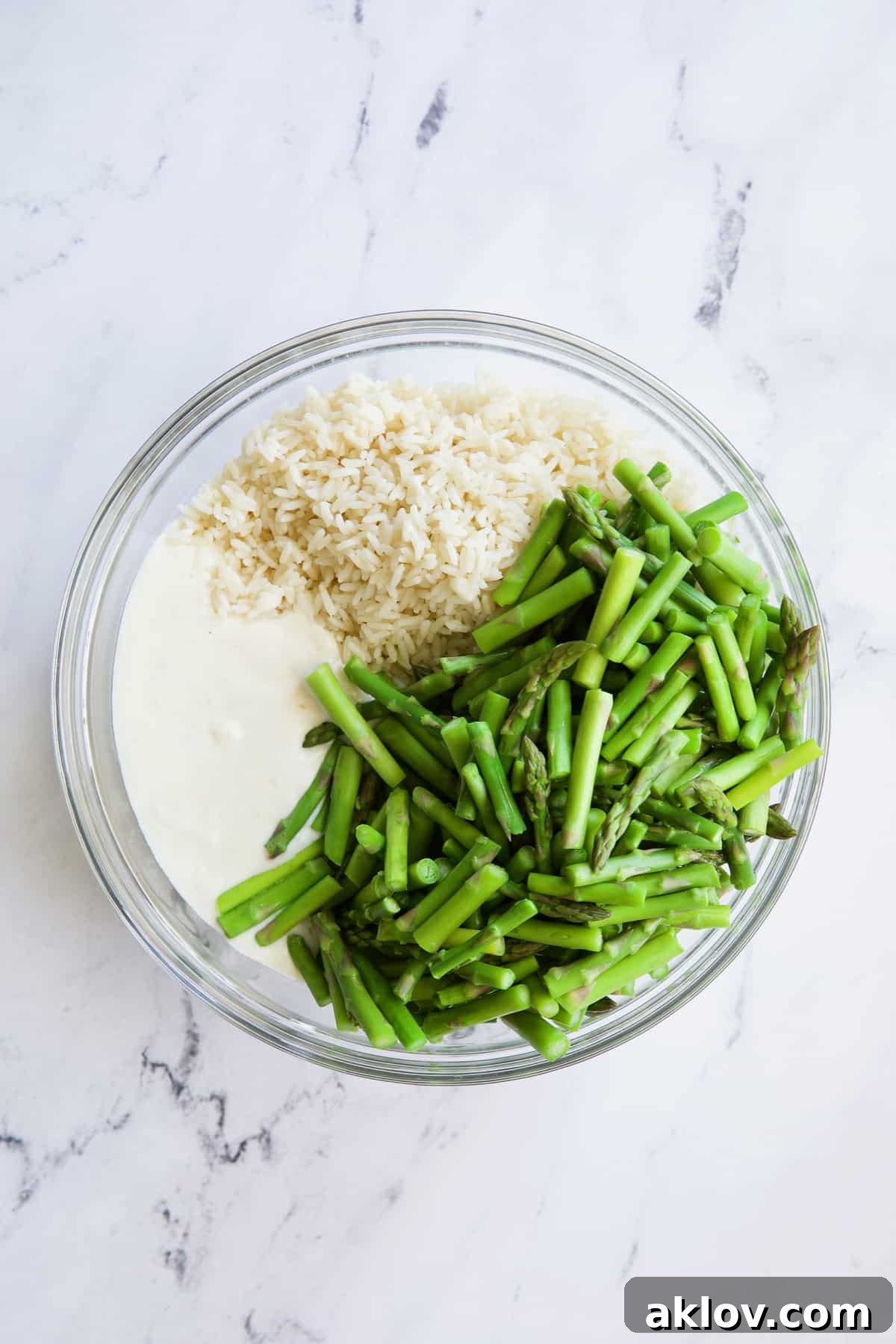 A large glass mixing bowl holds the cooked rice, blanched asparagus pieces, and the creamy sauce, expertly combined before transferring to the baking dish.