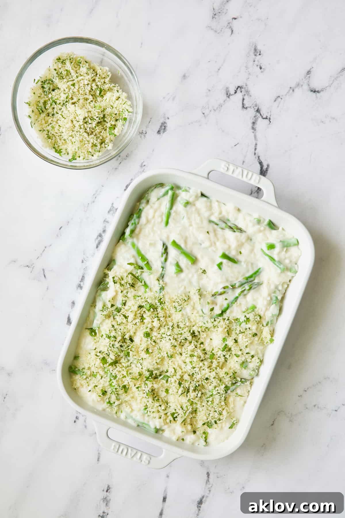 A hand gently sprinkles the golden breadcrumb topping over the creamy asparagus and rice casserole in a white baking dish, preparing it for the oven.