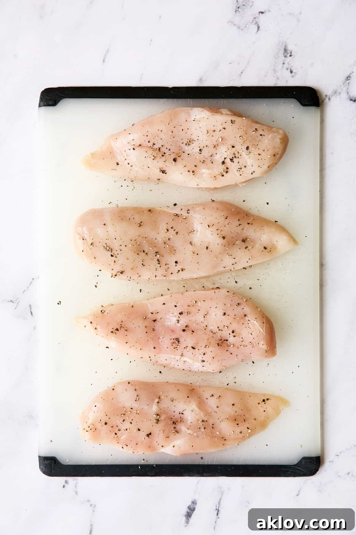 Four boneless skinless chicken breasts on a white cutting board seasoned with salt and pepper.
