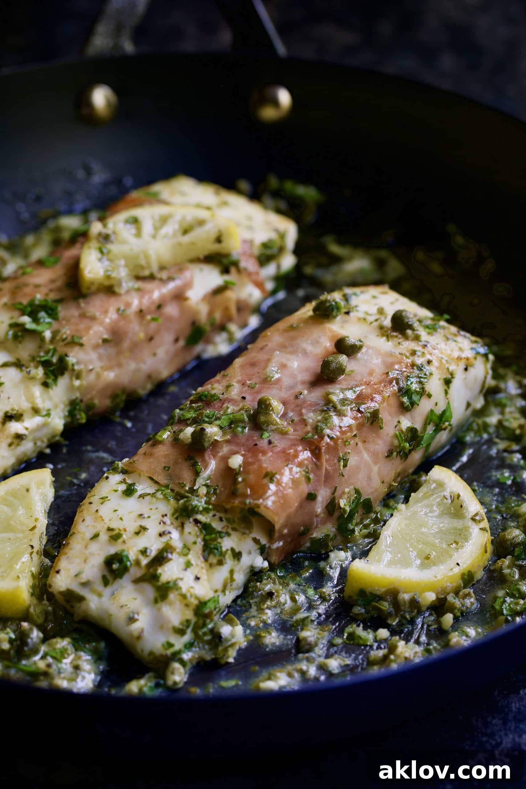 Prosciutto-Wrapped Halibut with Herb - Caper Butter in black skillet with lemon wedges.