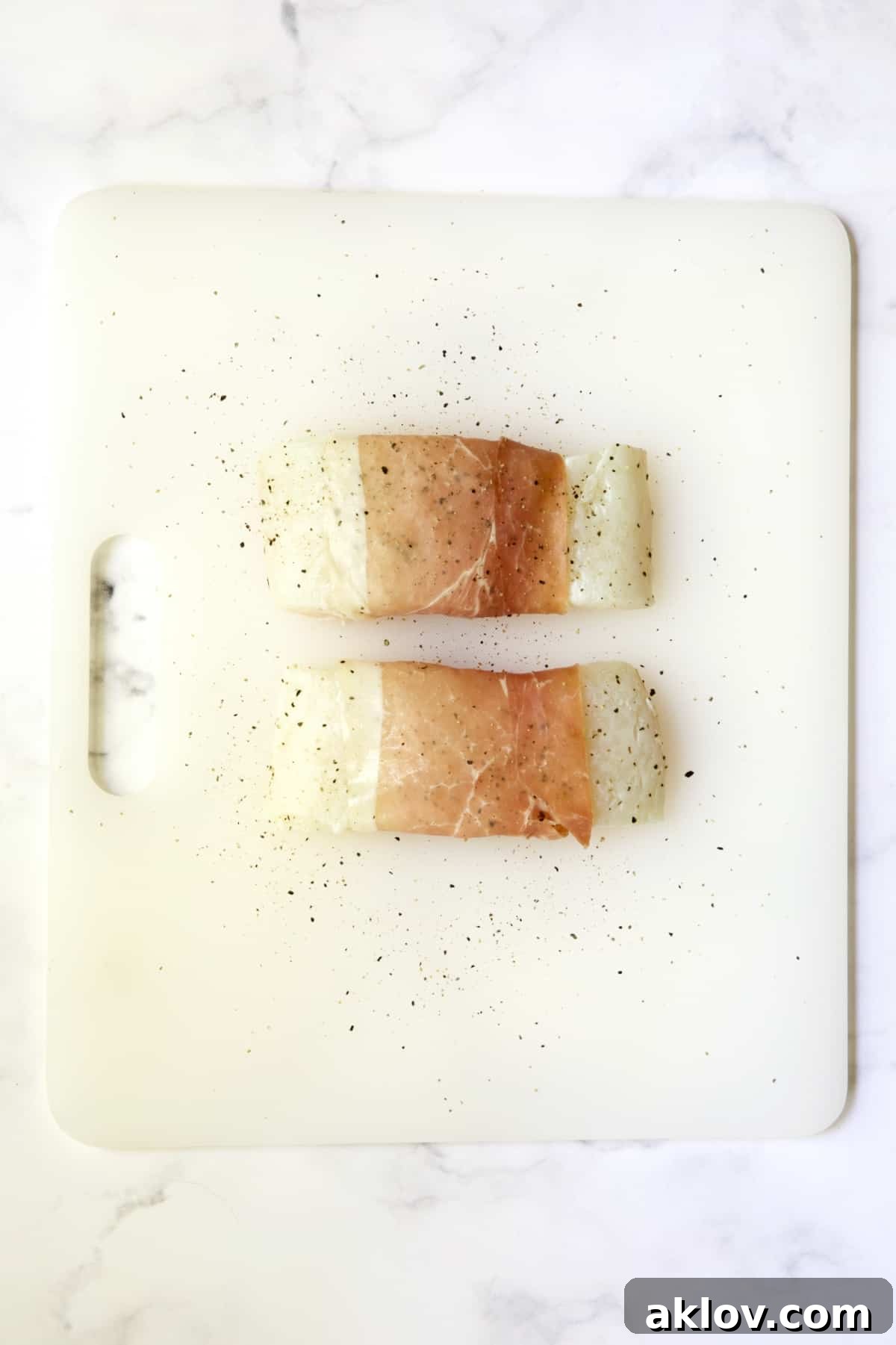 Two prosciutto-wrapped halibut fillets seasoned with pepper on a white plastic cutting board before being cooked.