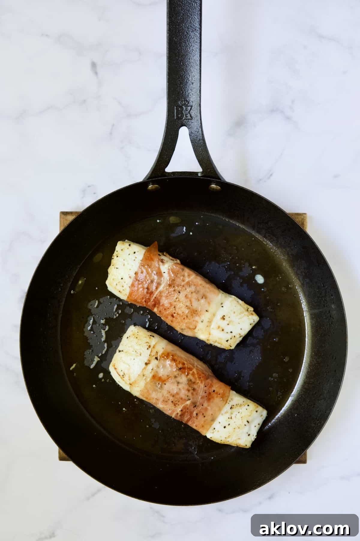 Two prosciutto-wrapped halibut fillets in black skillet being browned.