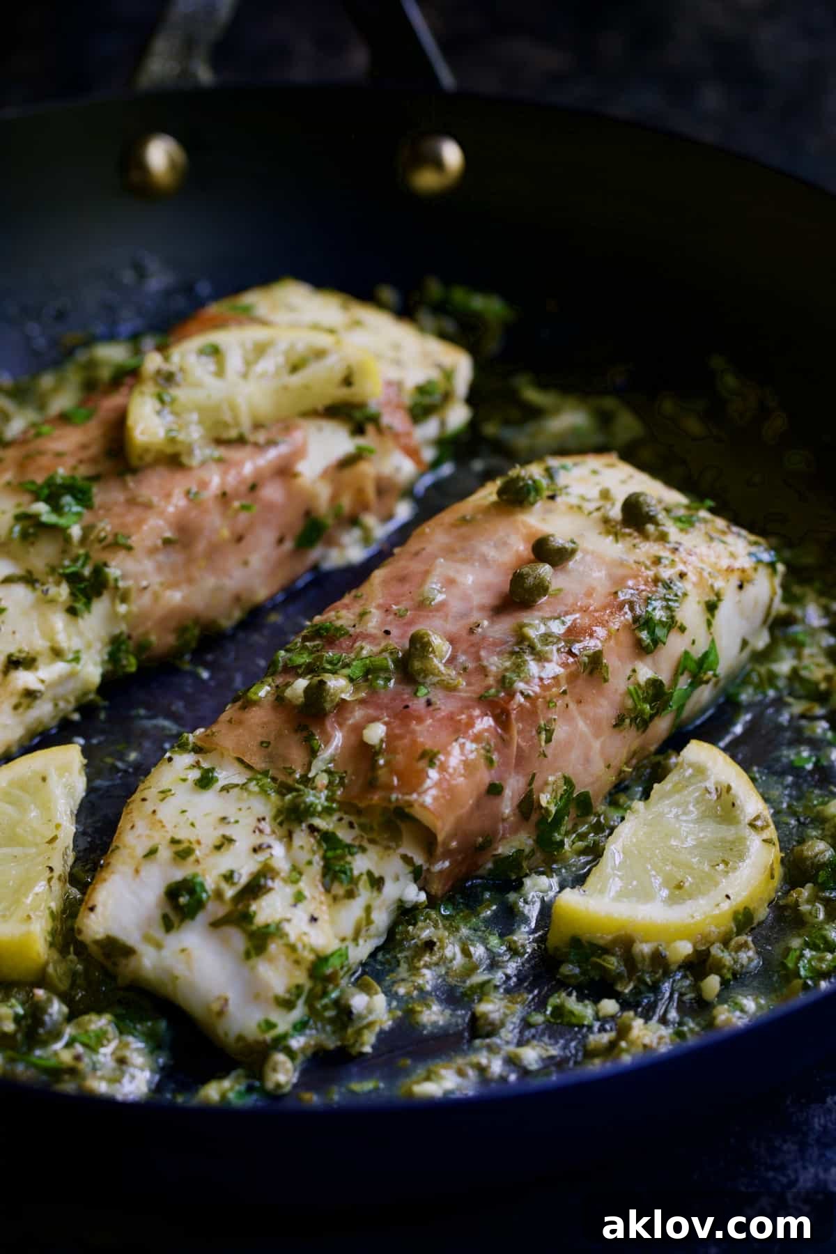 Prosciutto-Wrapped Halibut with Herb - Caper Butter with lemon wedges in black skillet.