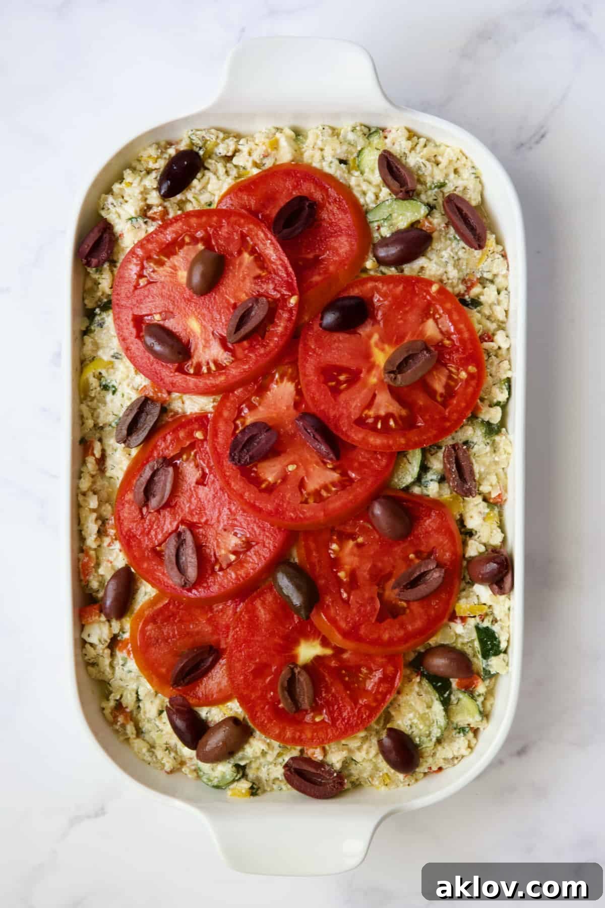 Assembled Greek Brown Rice and Vegetable Casserole, topped with sliced tomatoes and Kalamata olives, ready for baking.