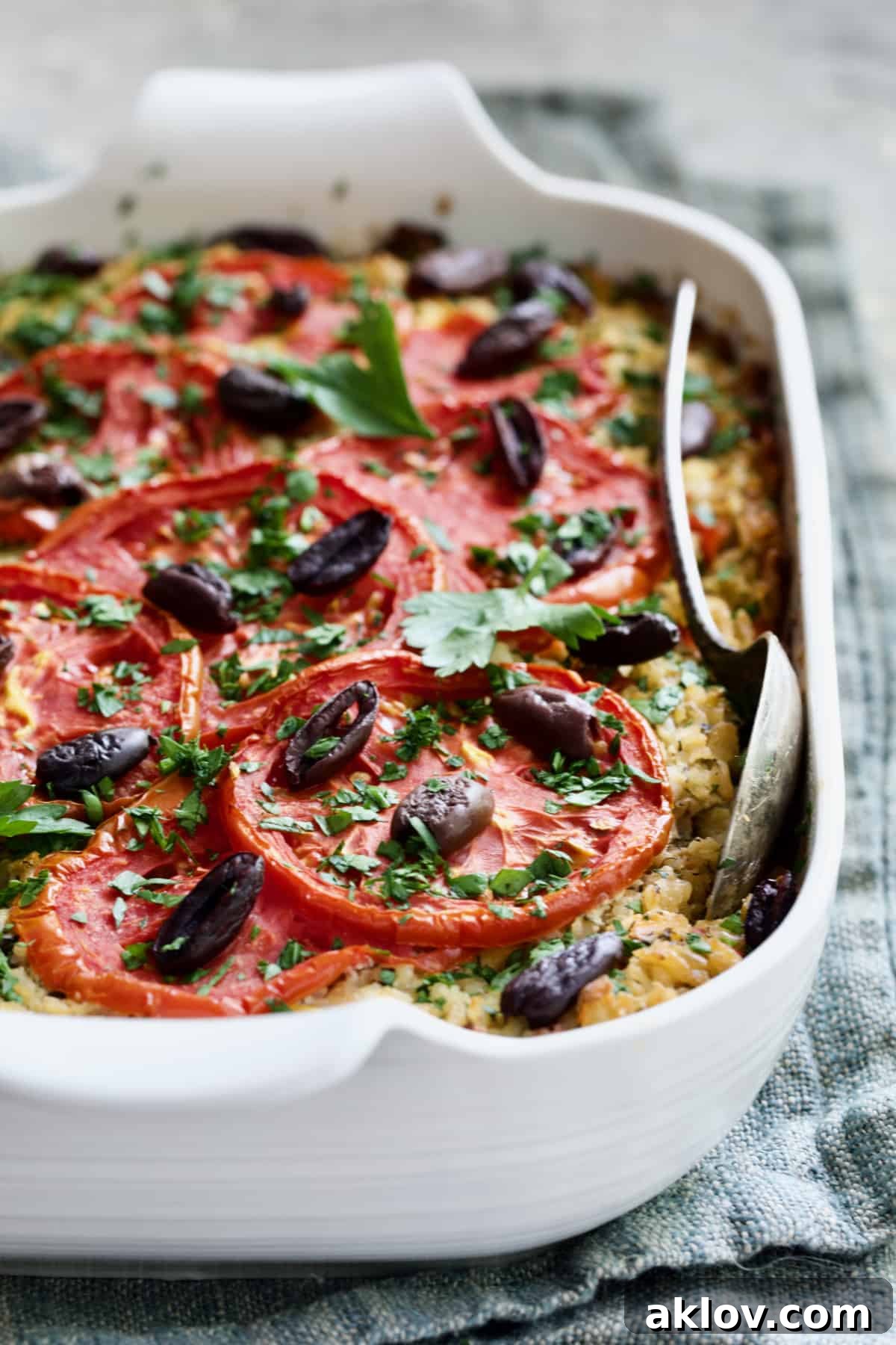 A generous serving of Greek Brown Rice and Vegetable Casserole in a white baking dish, with a serving spoon resting on a blue napkin, highlighting its rich, textured layers.