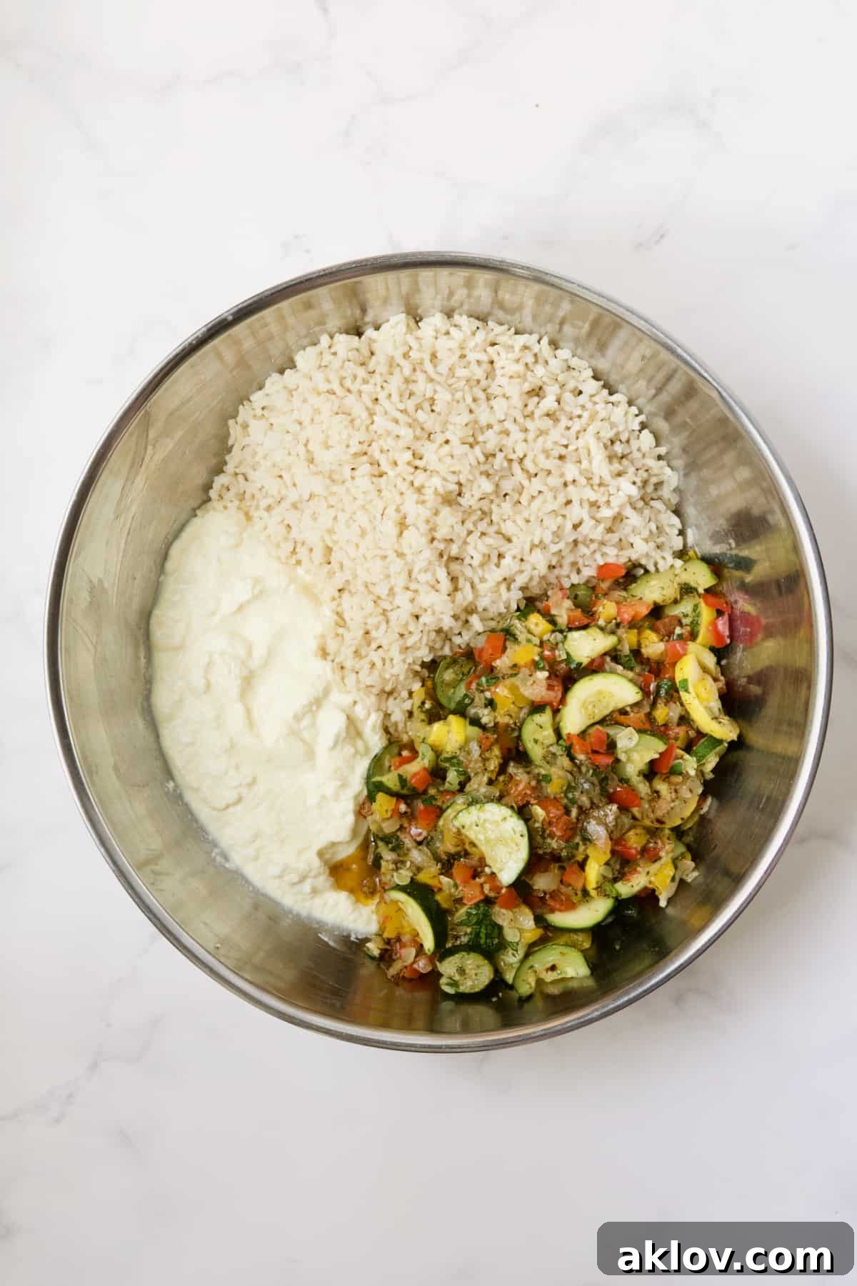 Separate ingredients for Greek Brown Rice and Vegetable Casserole in a stainless steel bowl, prior to mixing, showing vegetables, rice, and cheeses.