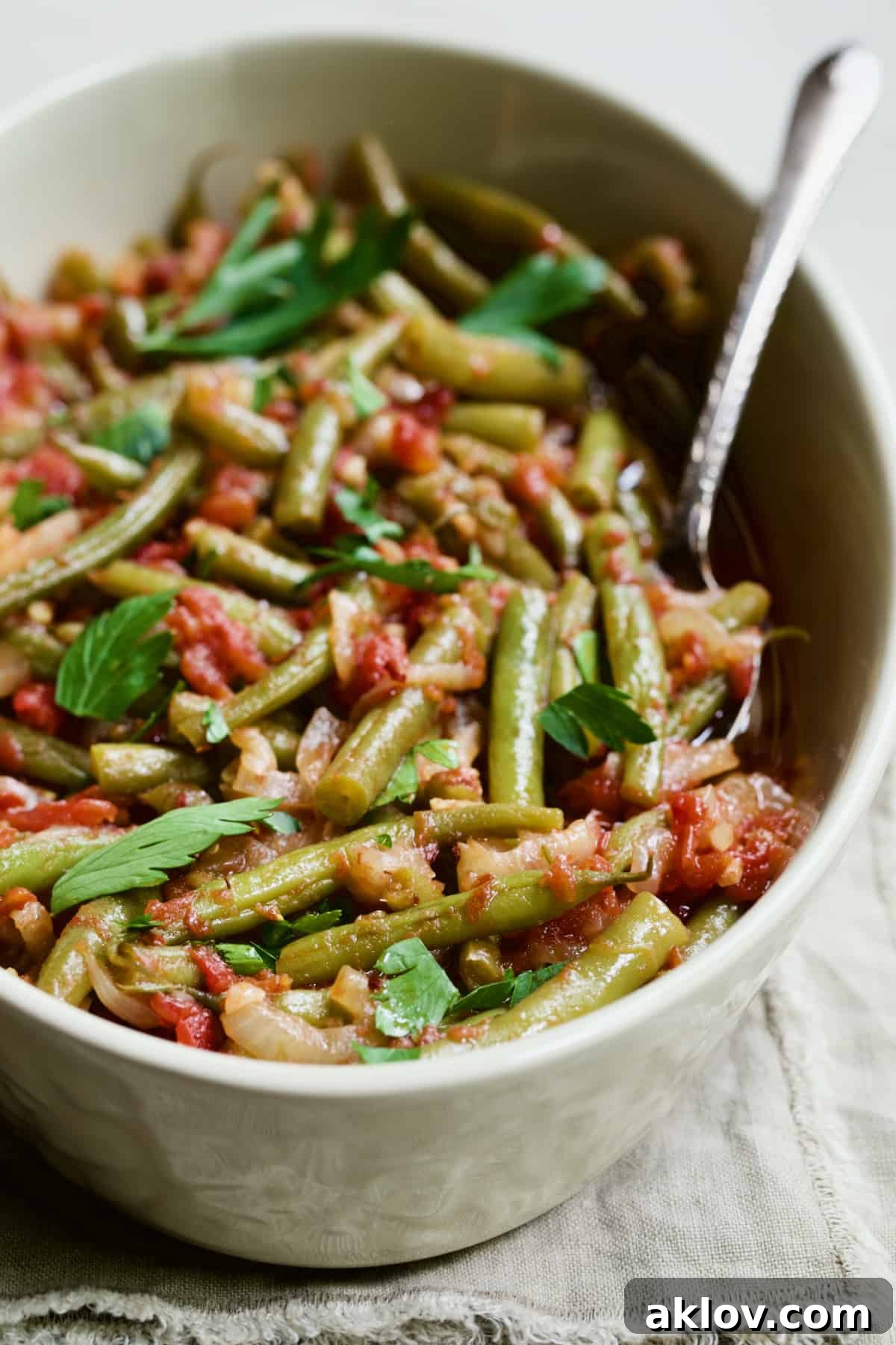 Mediterranean Green Bean and Tomato Stew 3 Mediterranean Braised Green Beans with Tomatoes in serving dish with serving spoon on beige fringed napkin.
