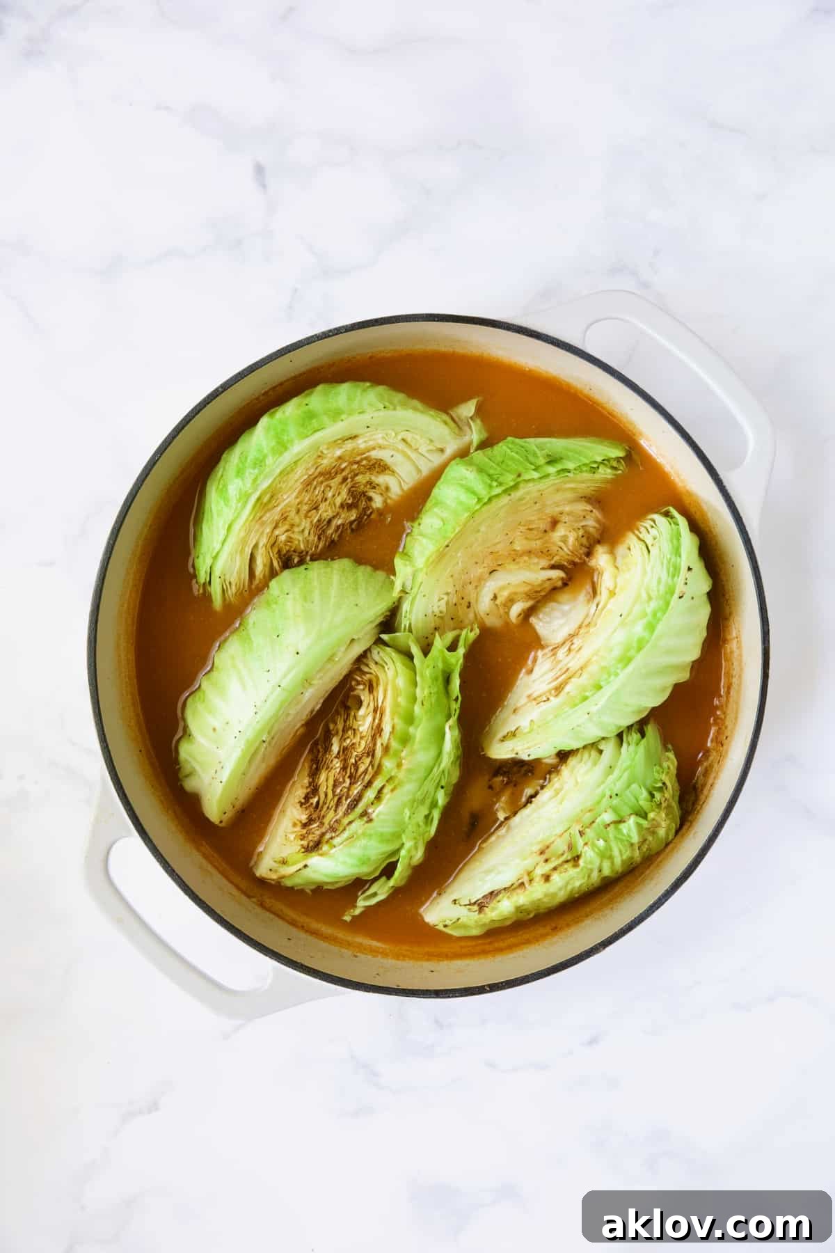 Browned cabbage wedges carefully placed back into the rich, simmering sauce within a white enamel cast iron skillet.
