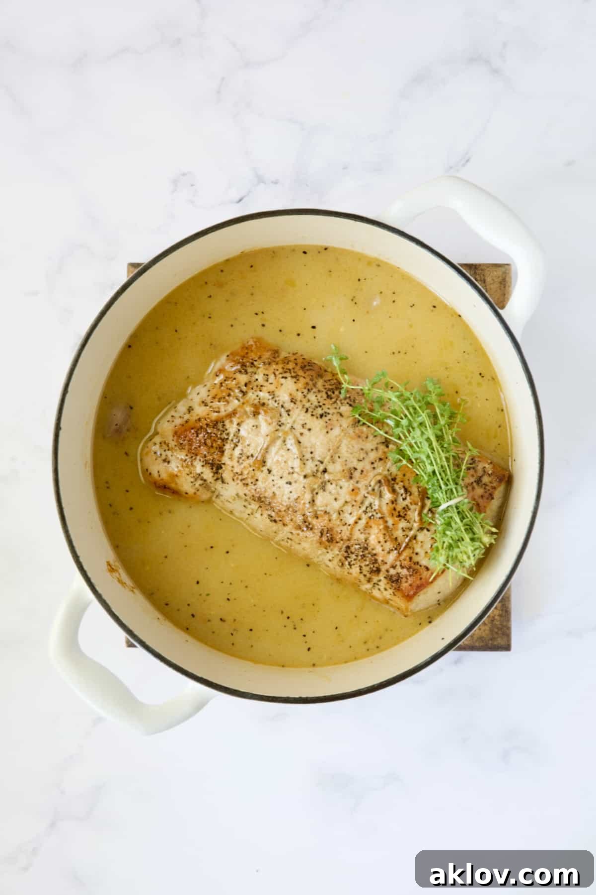 A pork loin roast nestled in a white Dutch oven, surrounded by the savory Dijon mustard braising liquid and fresh thyme sprigs, ready for the oven.