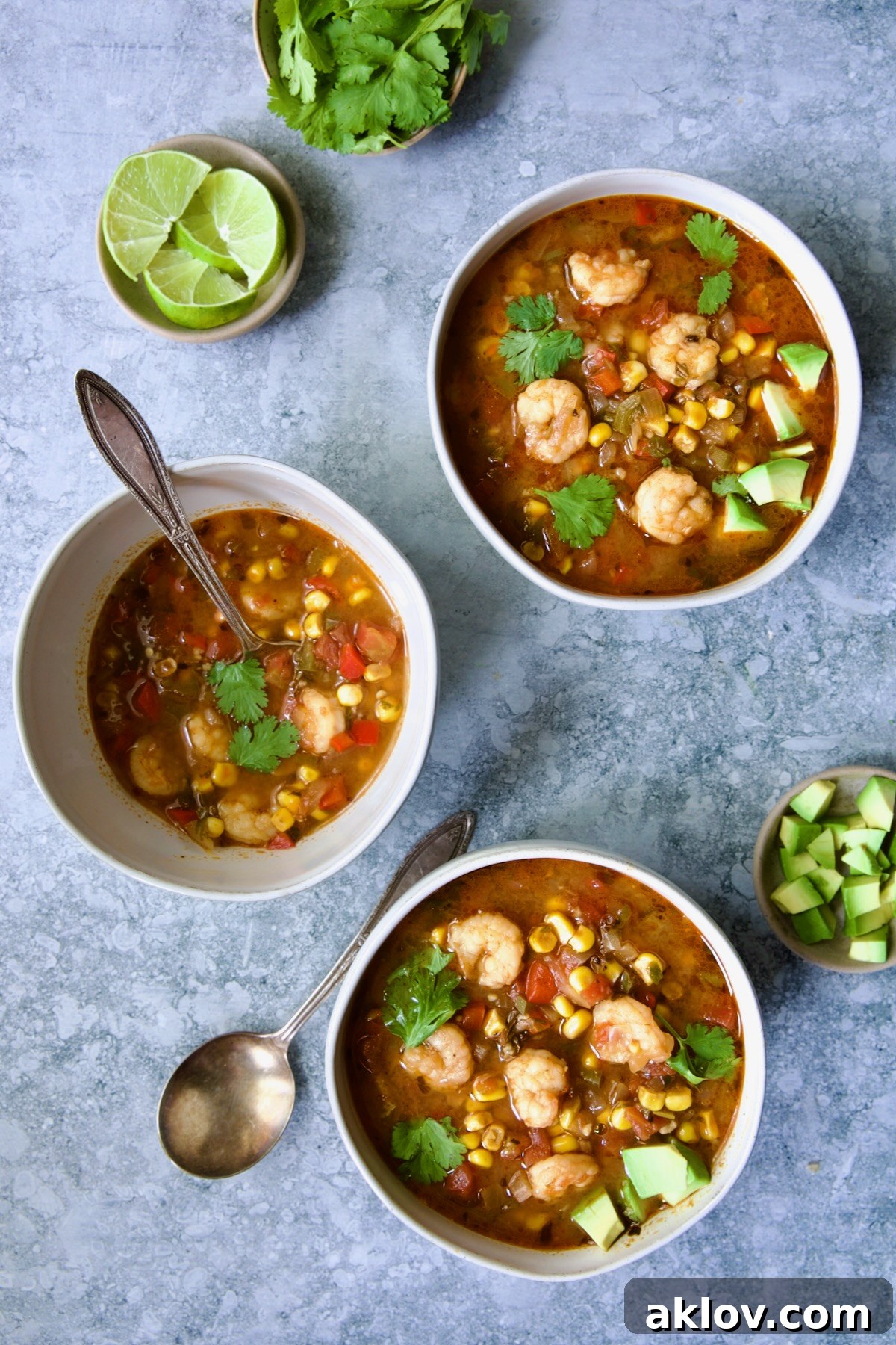 Three elegant bowls of Shrimp and Corn Soup, beautifully arranged with smaller bowls of fresh lime wedges, chopped cilantro, and diced avocado for garnishing.