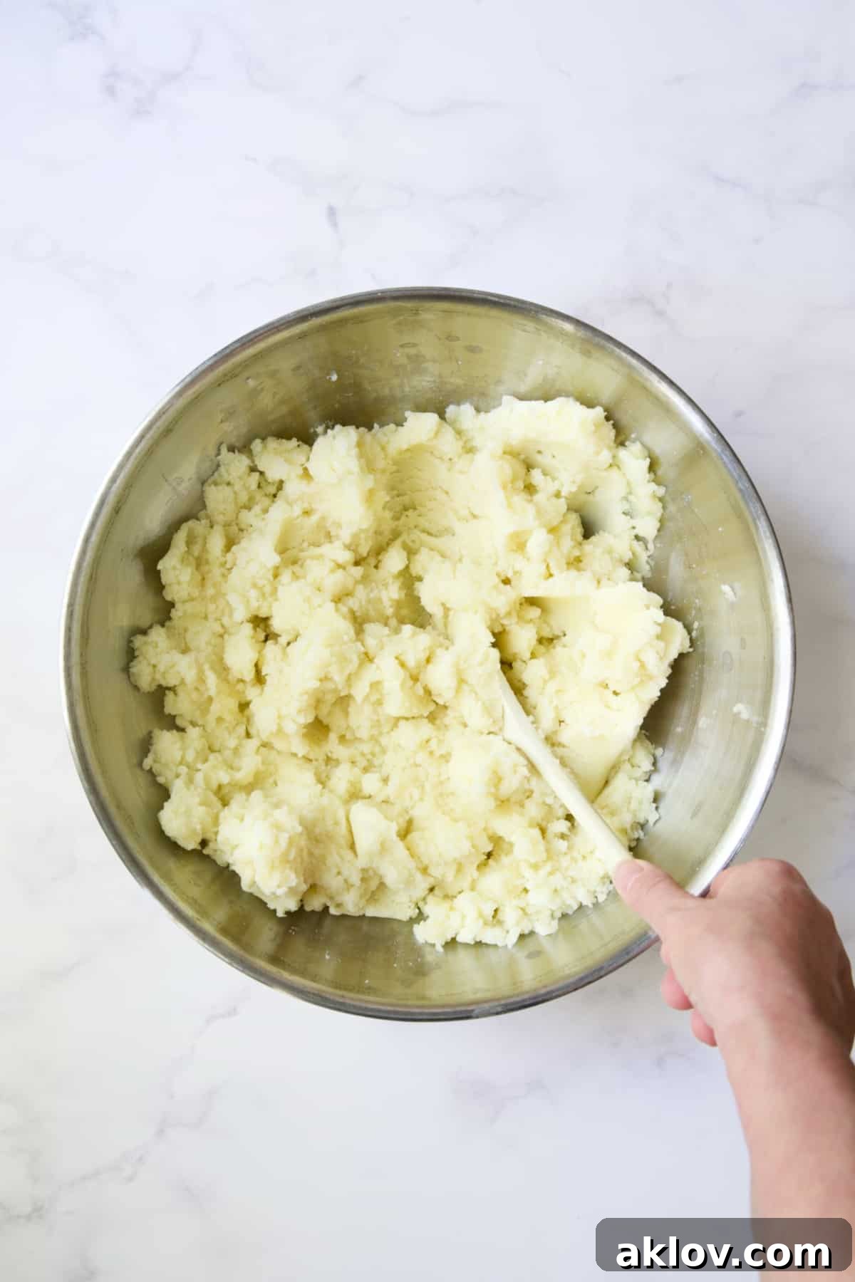 Freshly riced potatoes being gently stirred in a stainless steel bowl, ensuring the melted butter is evenly distributed for a rich, smooth texture.