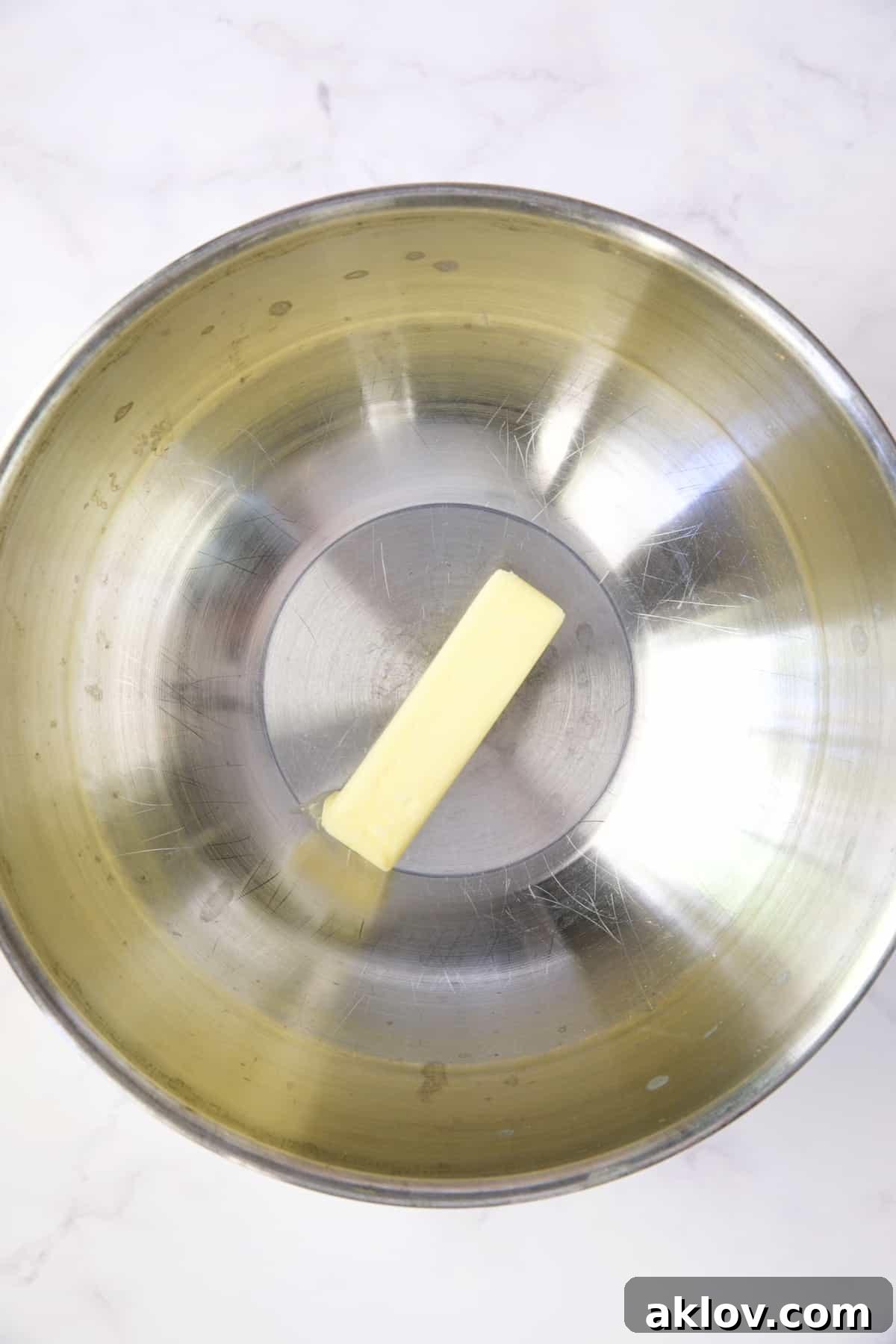 A stick of unsalted butter resting in a stainless steel bowl positioned over a pot on a stovetop.