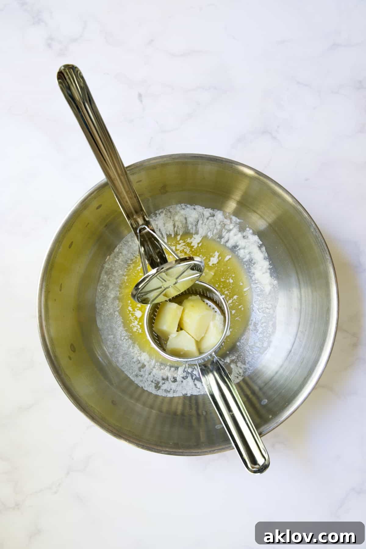 A potato ricer pressing cooked potatoes and garlic into a bowl of melted butter, creating light, fluffy strands.