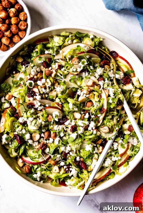 Shaved Brussels Sprouts Salad in white bowl with serving utensils, featuring apples and goat cheese.