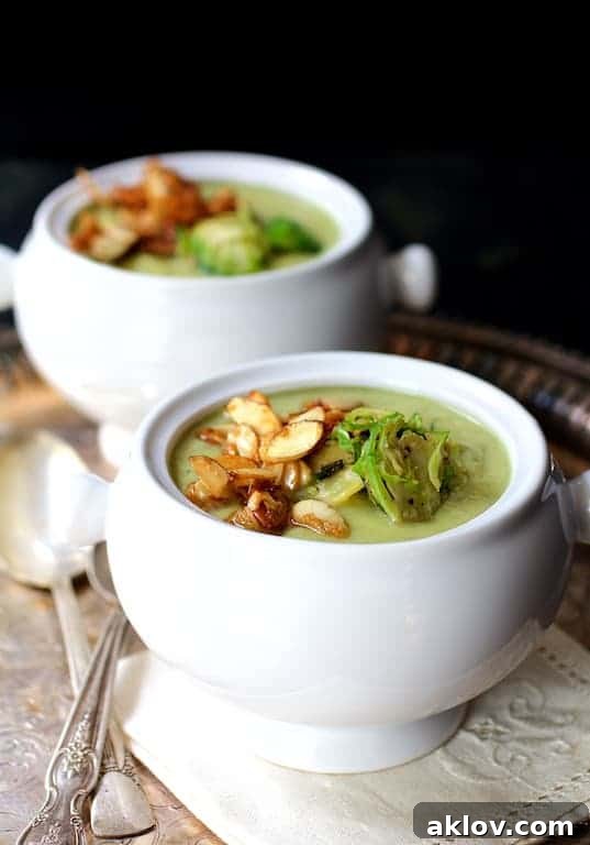 Two bowls of Cream of Brussels Sprouts Soup on a decorative napkin with soup spoons on a silver tray, garnished with toasted almonds.