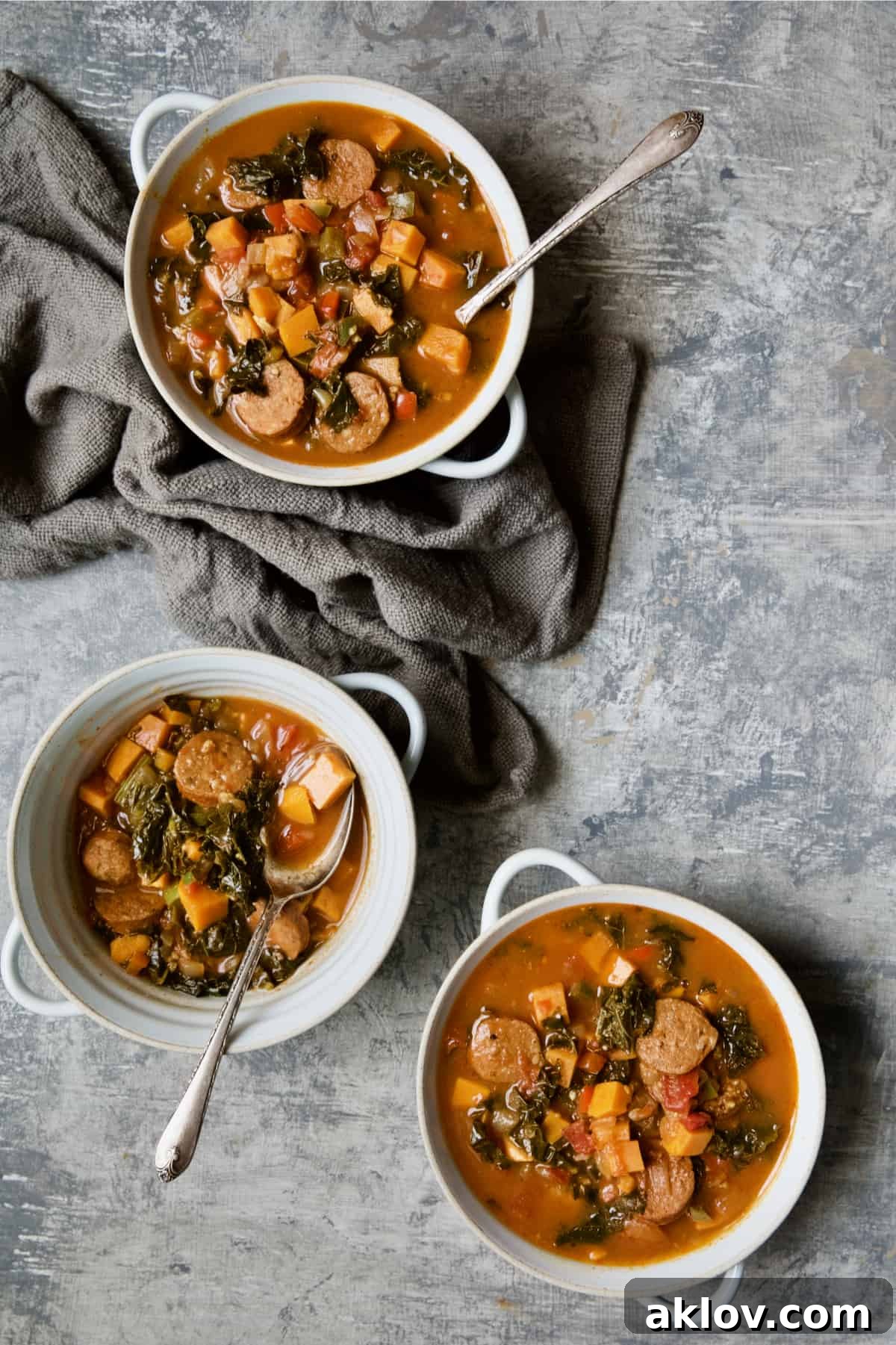 Three bowls of Chorizo Sweet Potato Stew with Kale on gray background with gray napkin.
