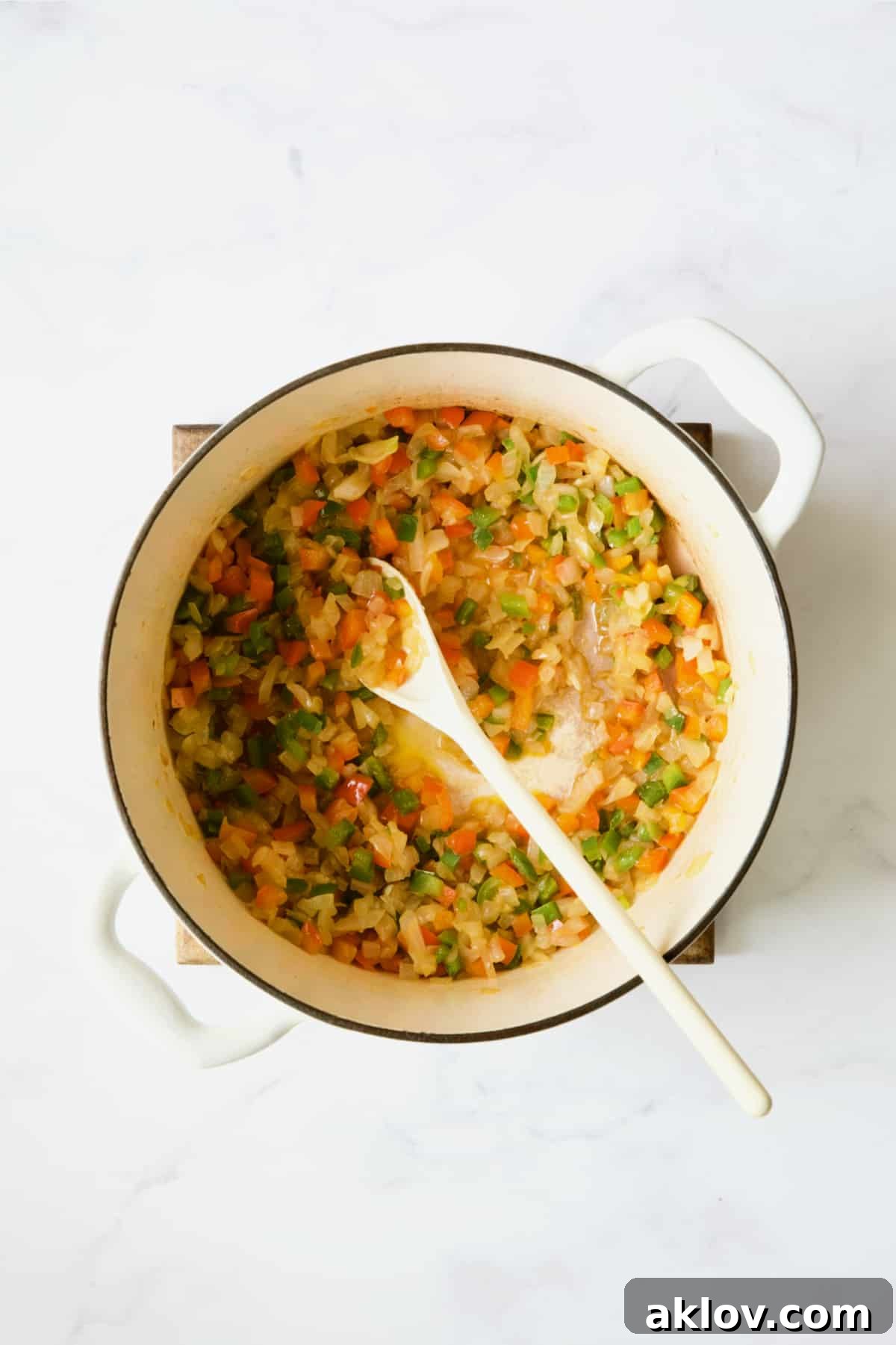Onions and peppers being cooked in white Dutch oven with white mixing spoon.