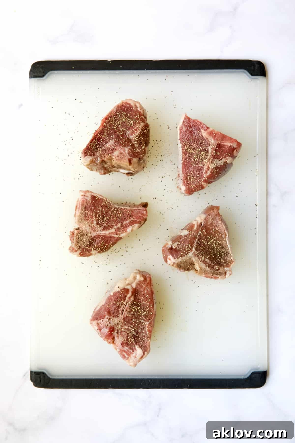 Five lamb chops on a white plastic cutting board, perfectly seasoned with salt and pepper.