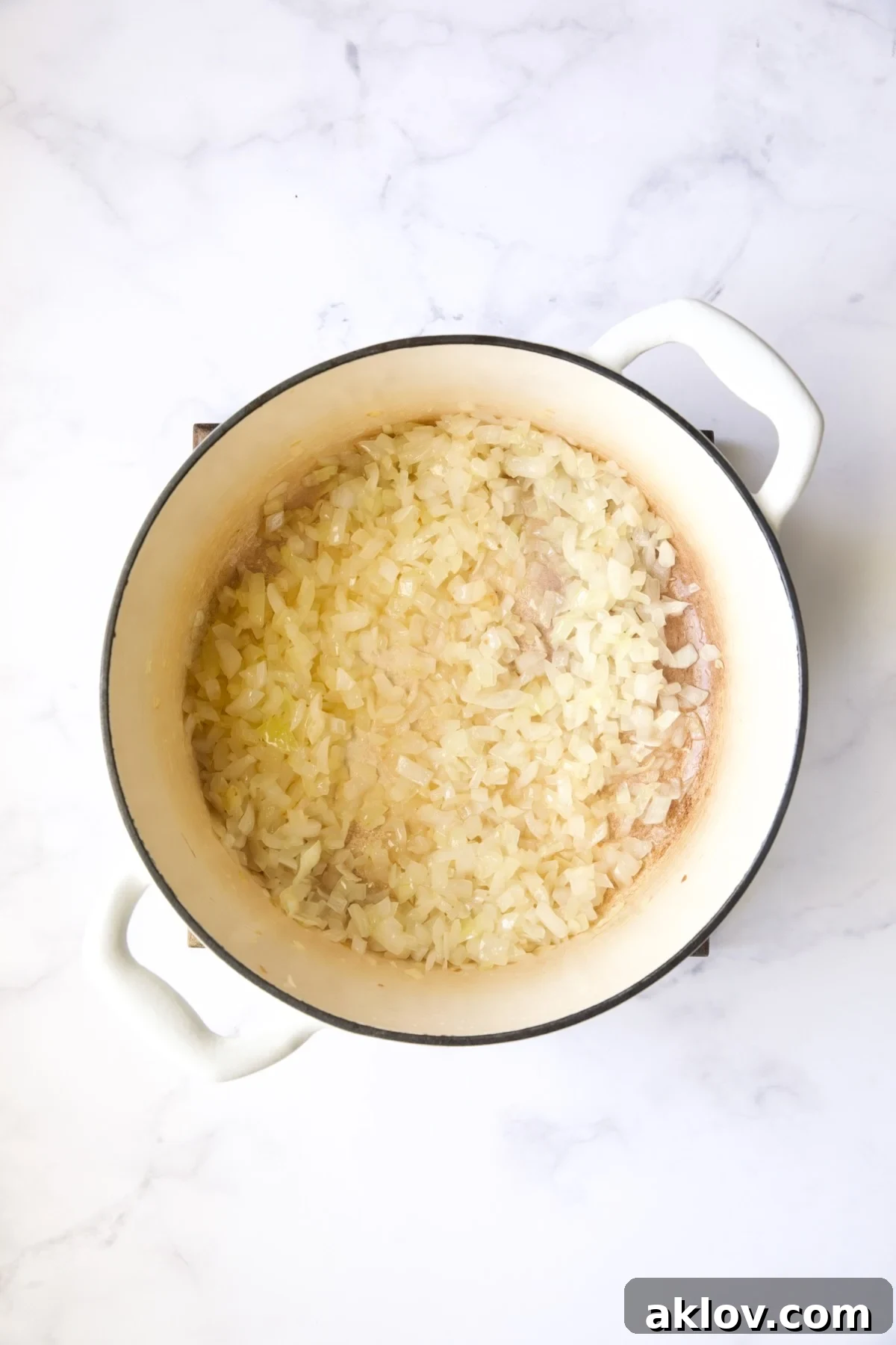Translucent, cooked onions simmering gently in a white Dutch oven, forming the fragrant aromatic base of the stew.