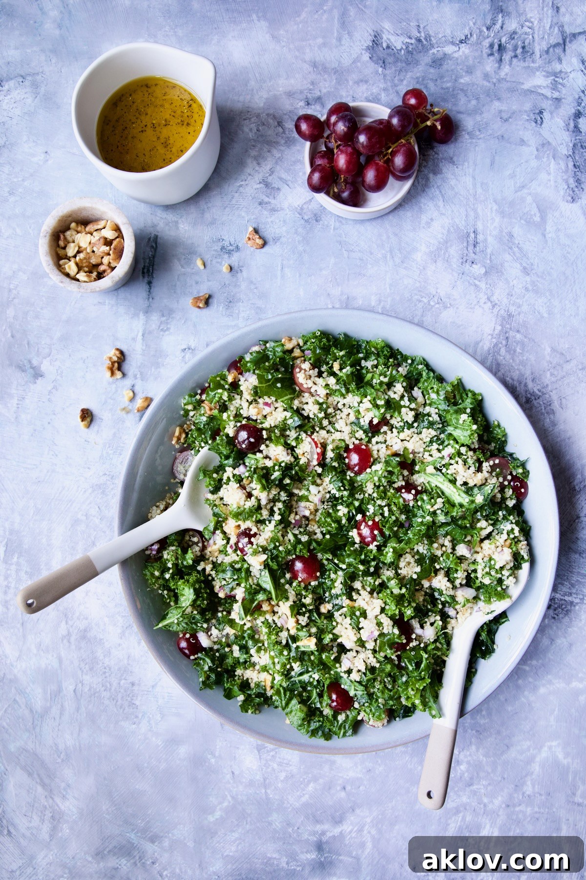 The finished Kale Quinoa Salad with Lemon Honey Dressing in a serving bowl, artfully arranged on a blue background, with separate bowls of walnuts, dressing, and grapes highlighting its fresh components.