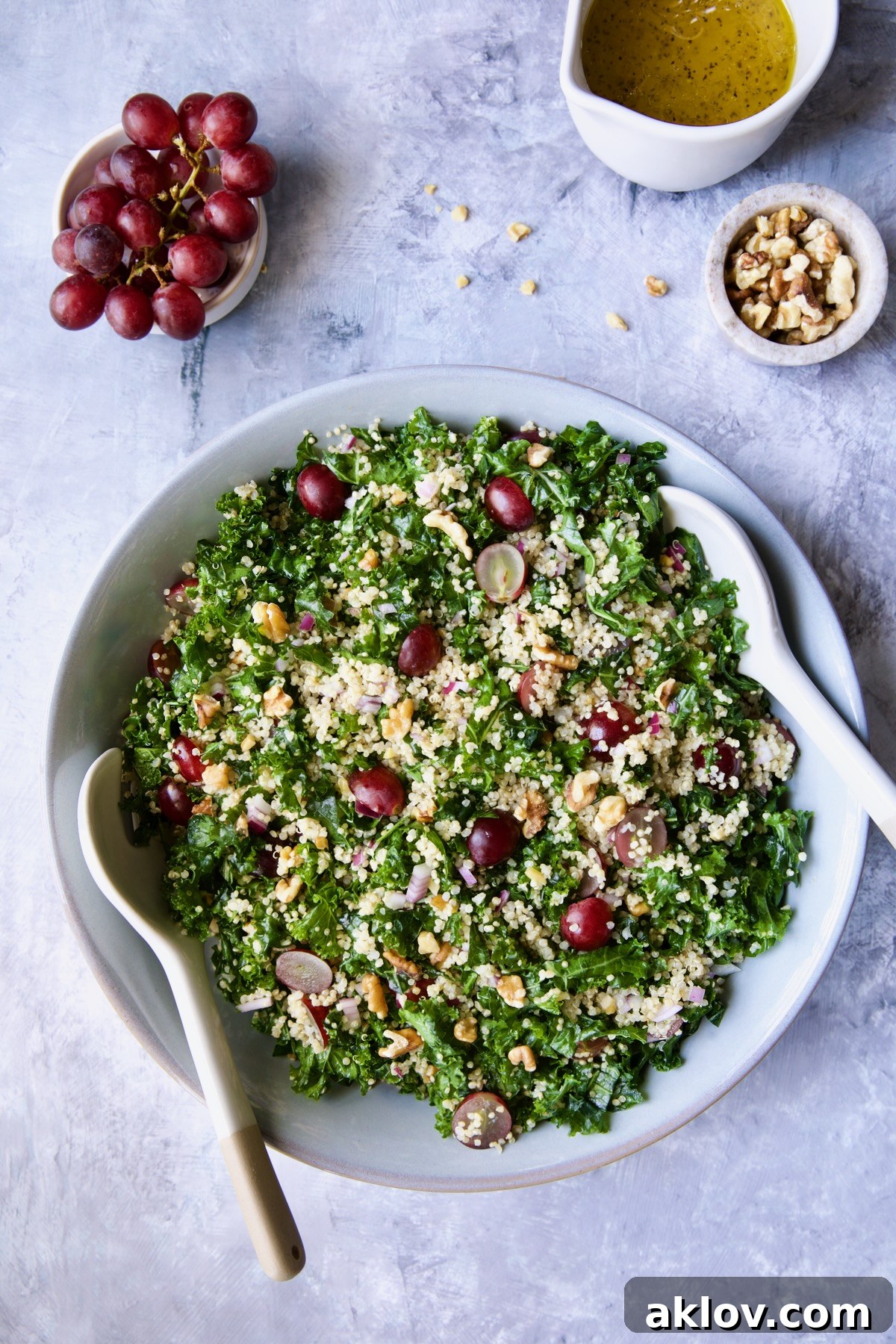 A comprehensive spread featuring the Kale Quinoa Salad with Lemon Honey Dressing in a gray serving bowl, complemented by separate bowls of grapes, dressing, and walnuts, highlighting all its fresh ingredients.