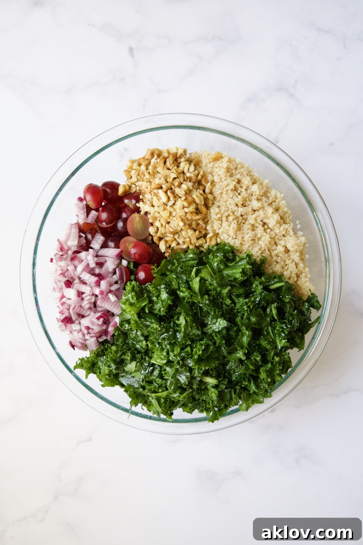 A vibrant mix of massaged kale, cooked quinoa, walnuts, red grapes, and red onion in a glass bowl, waiting to be tossed together.