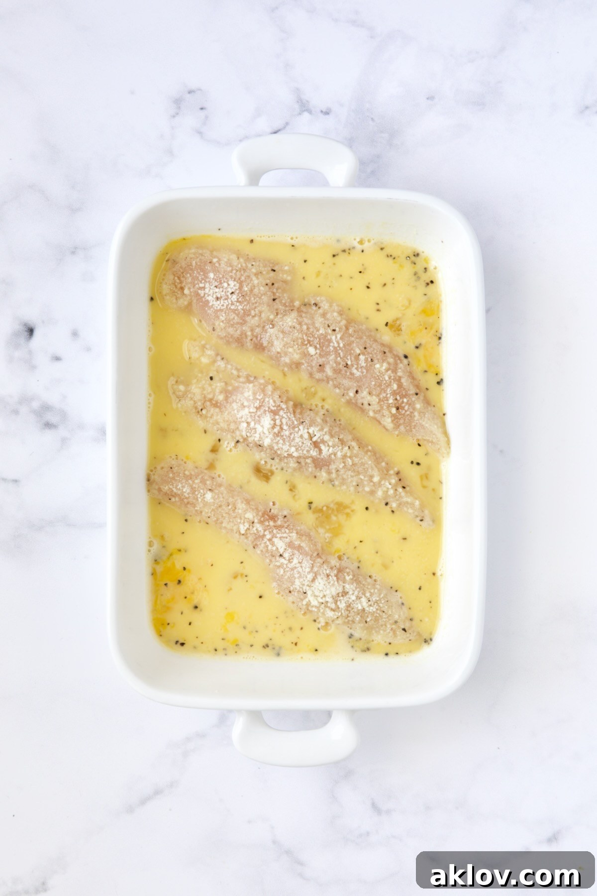 Three chicken tenders in beaten egg after being coated with almond flour in white rectangular baking pan.