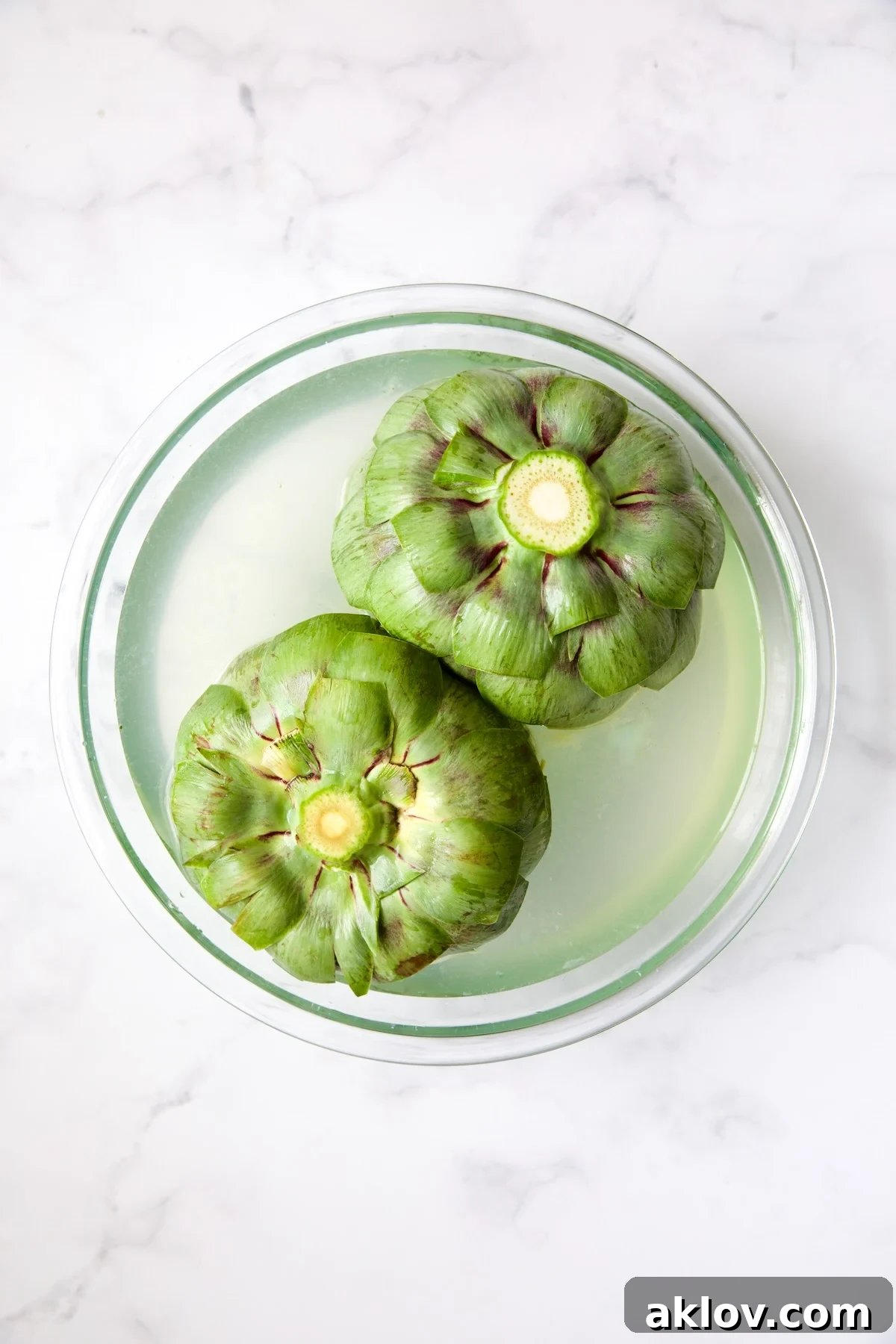 Savory Italian Stuffed Artichokes 12 Two uncooked artichokes submerged in lemon water in glass bowl.