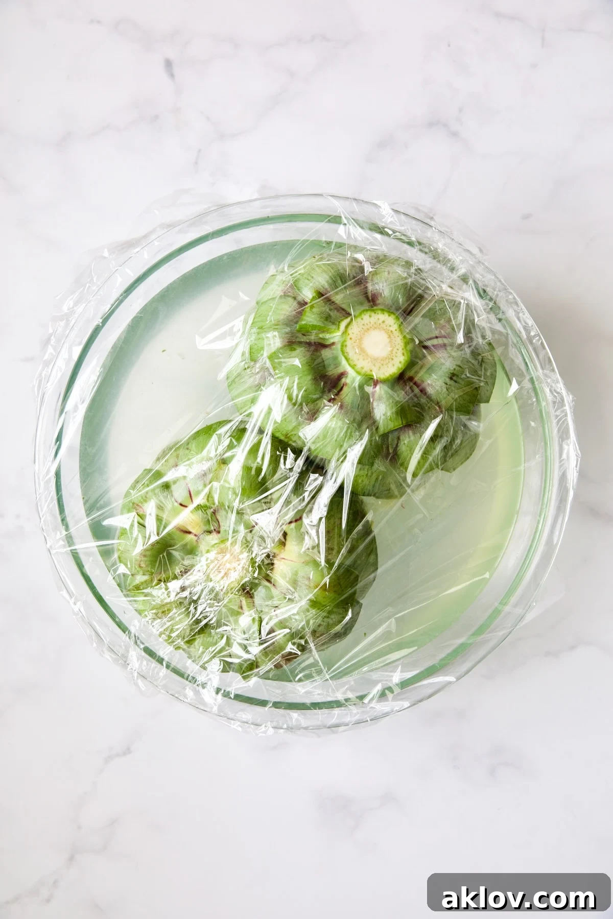Savory Italian Stuffed Artichokes 13 Two artichokes submerged in lemon water covered with plastic wrap.